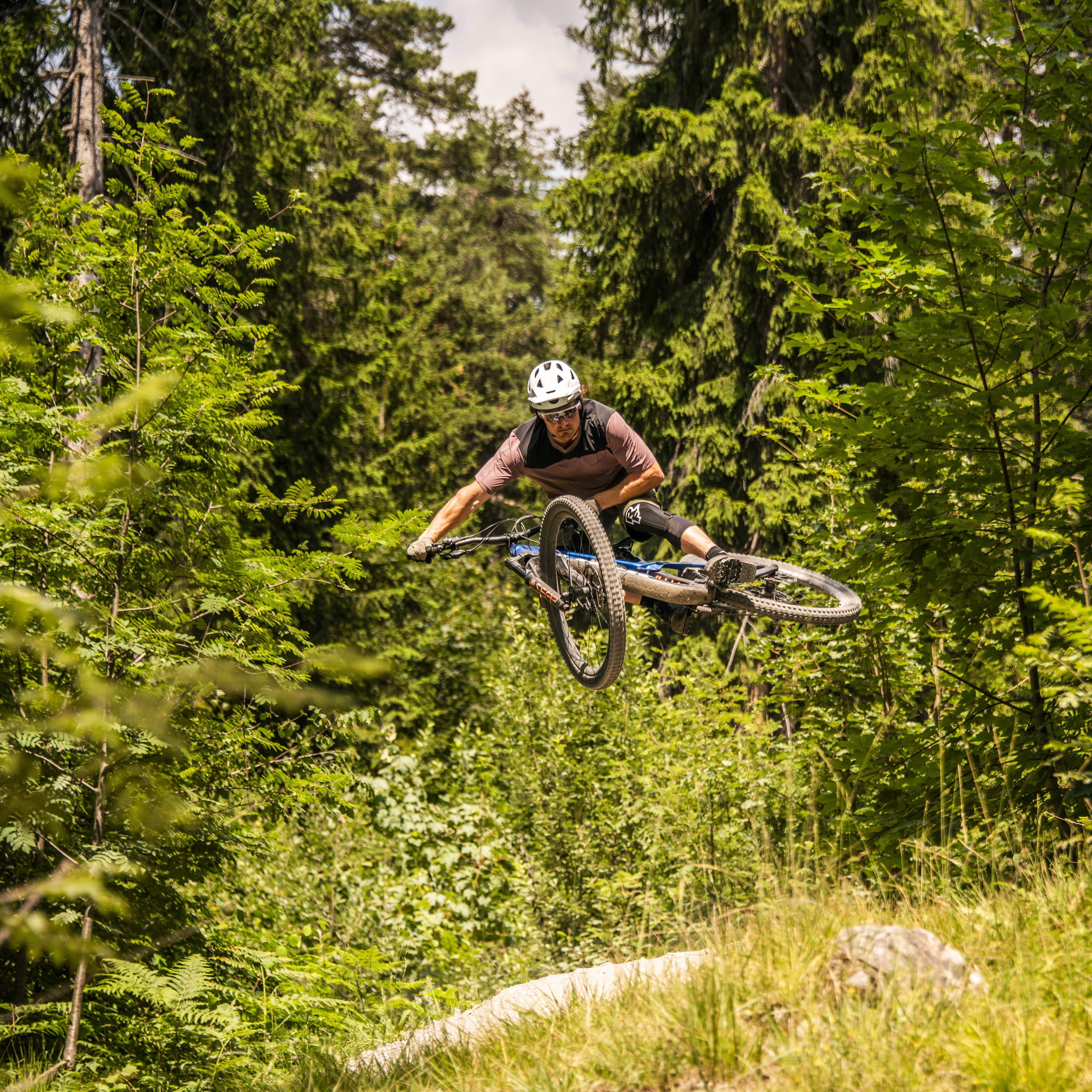 A man in a Norrøna outfit rides a mountain bike through a forest. He is mid-air over a jump.