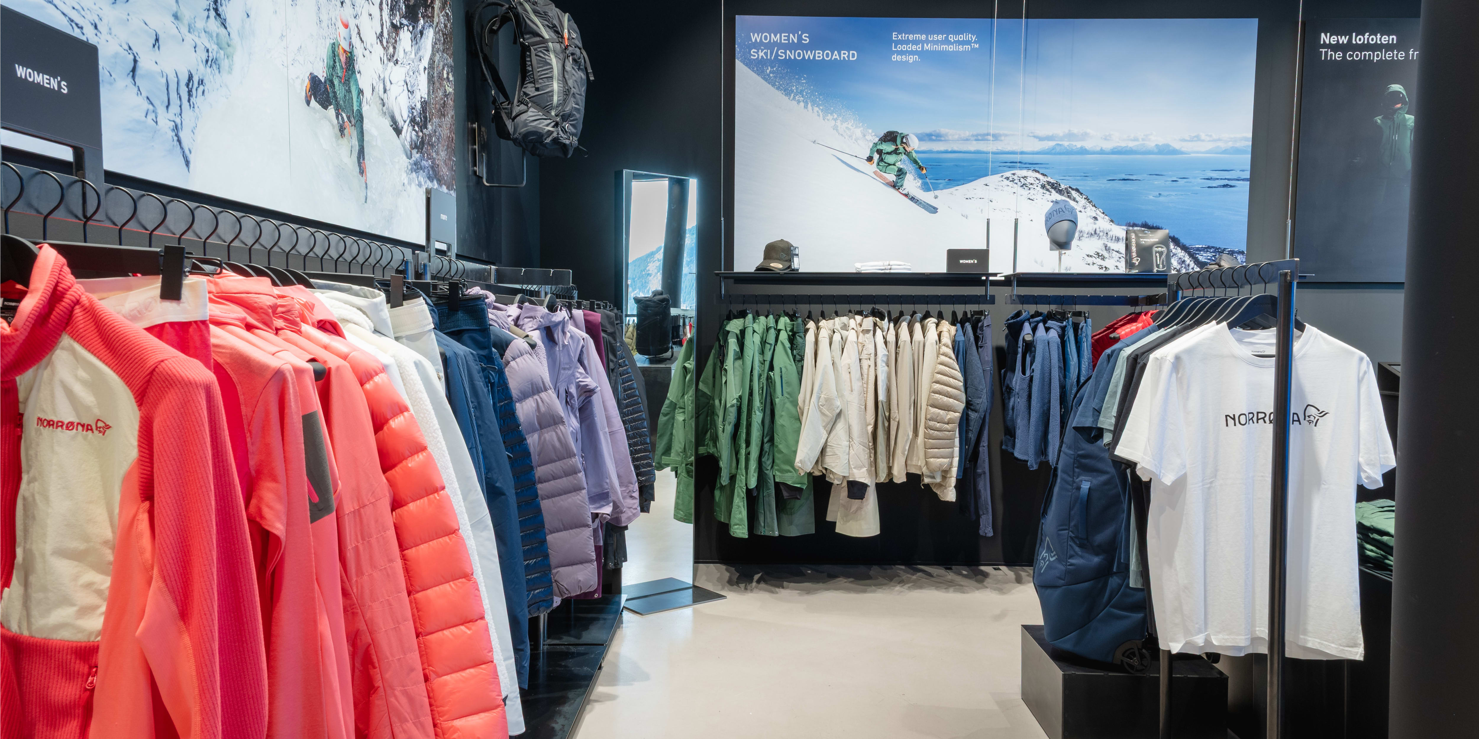 A store aisle filled with Norrøna clothing racks featuring jackets, shirts, and a backpack, against a backdrop of snowy mountain imagery.