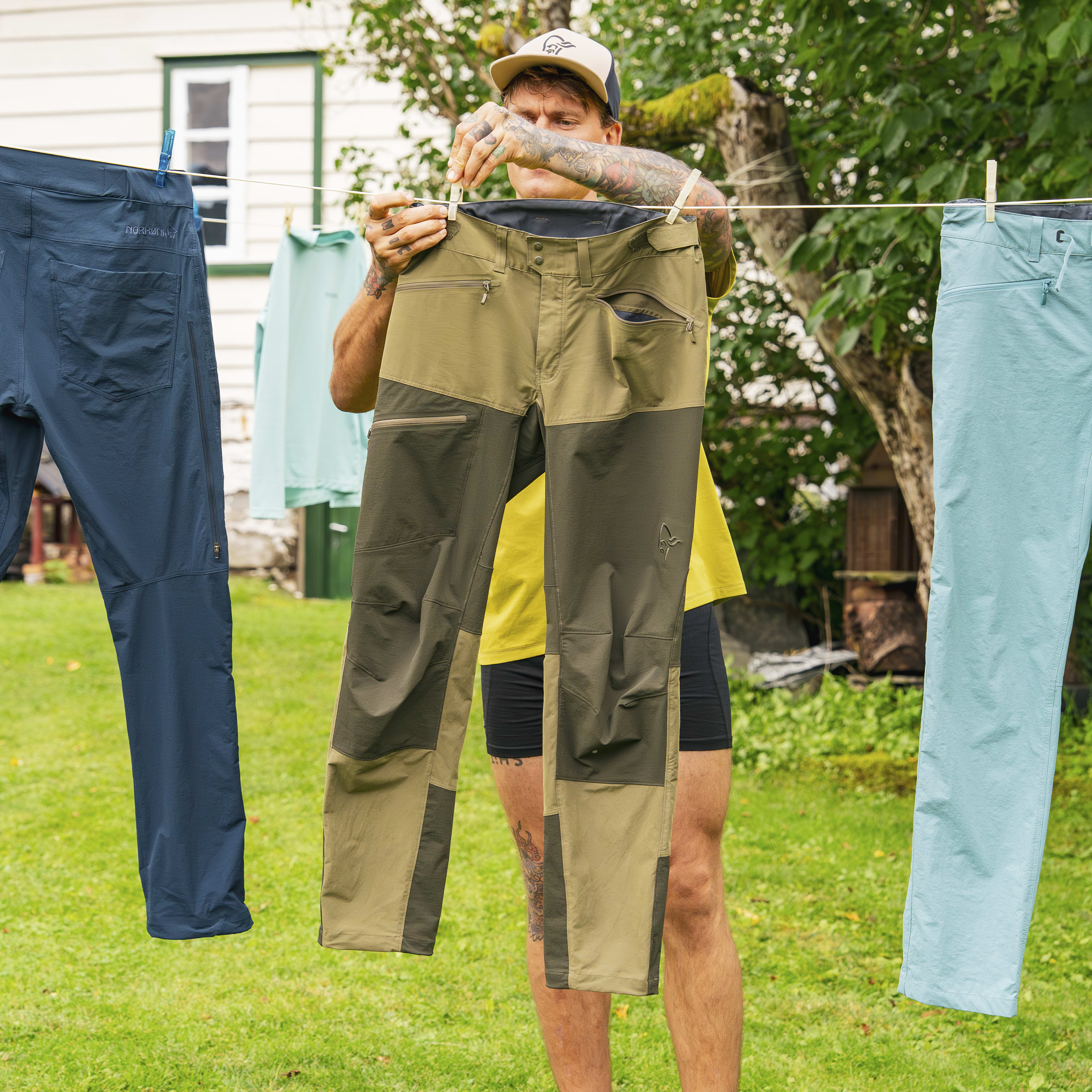 A tattooed man hangs Norrøna pants on a clothesline. Three pairs of Norrøna pants are displayed in varying colors.