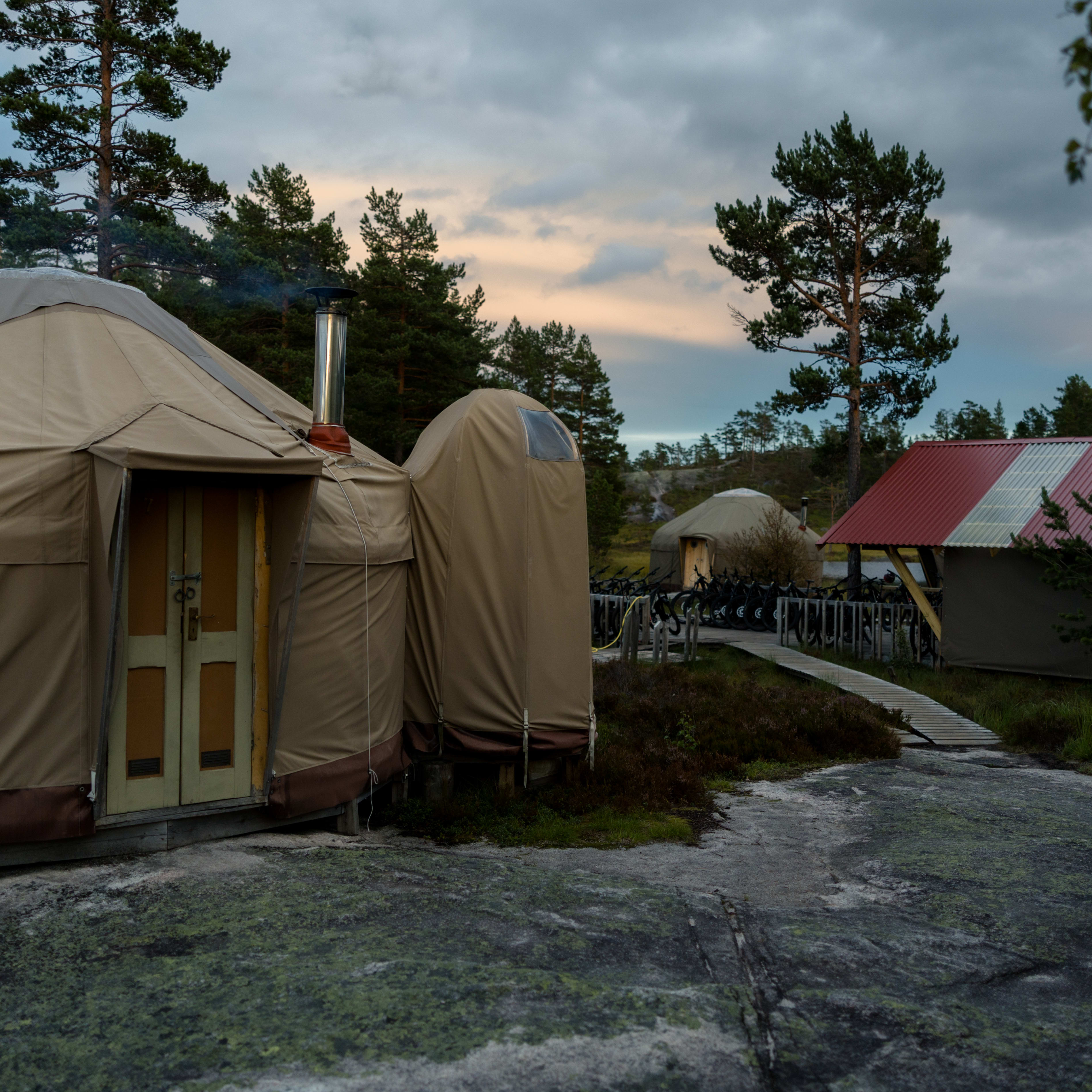 A tranquil outdoor scene shows yurts on a rocky landscape with trees, under a cloudy sky. One yurt features a chimney.
