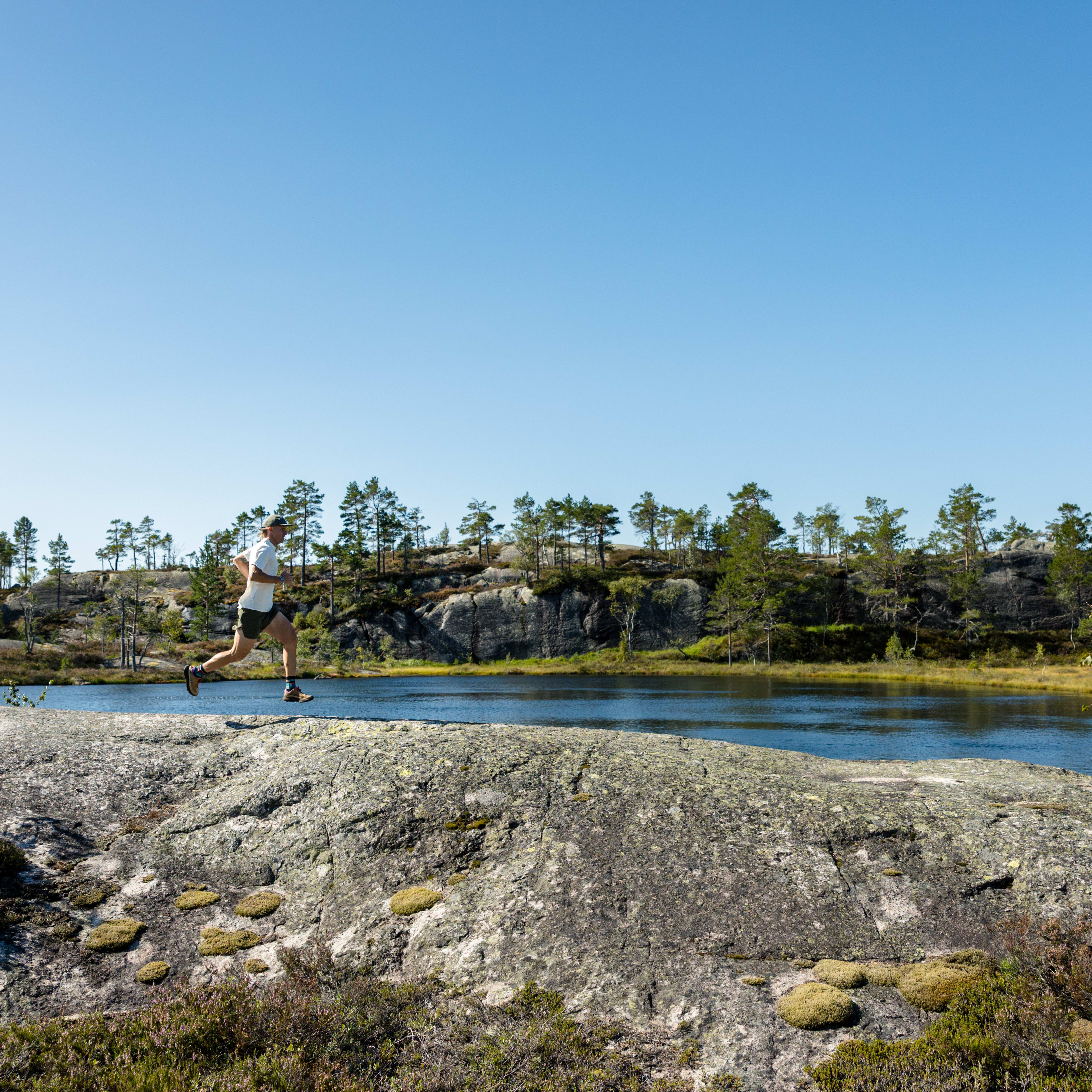 A man runs across a rocky outcrop next to a lake surrounded by trees under a clear blue sky. He wears Norrøna shorts and a white t-shirt.