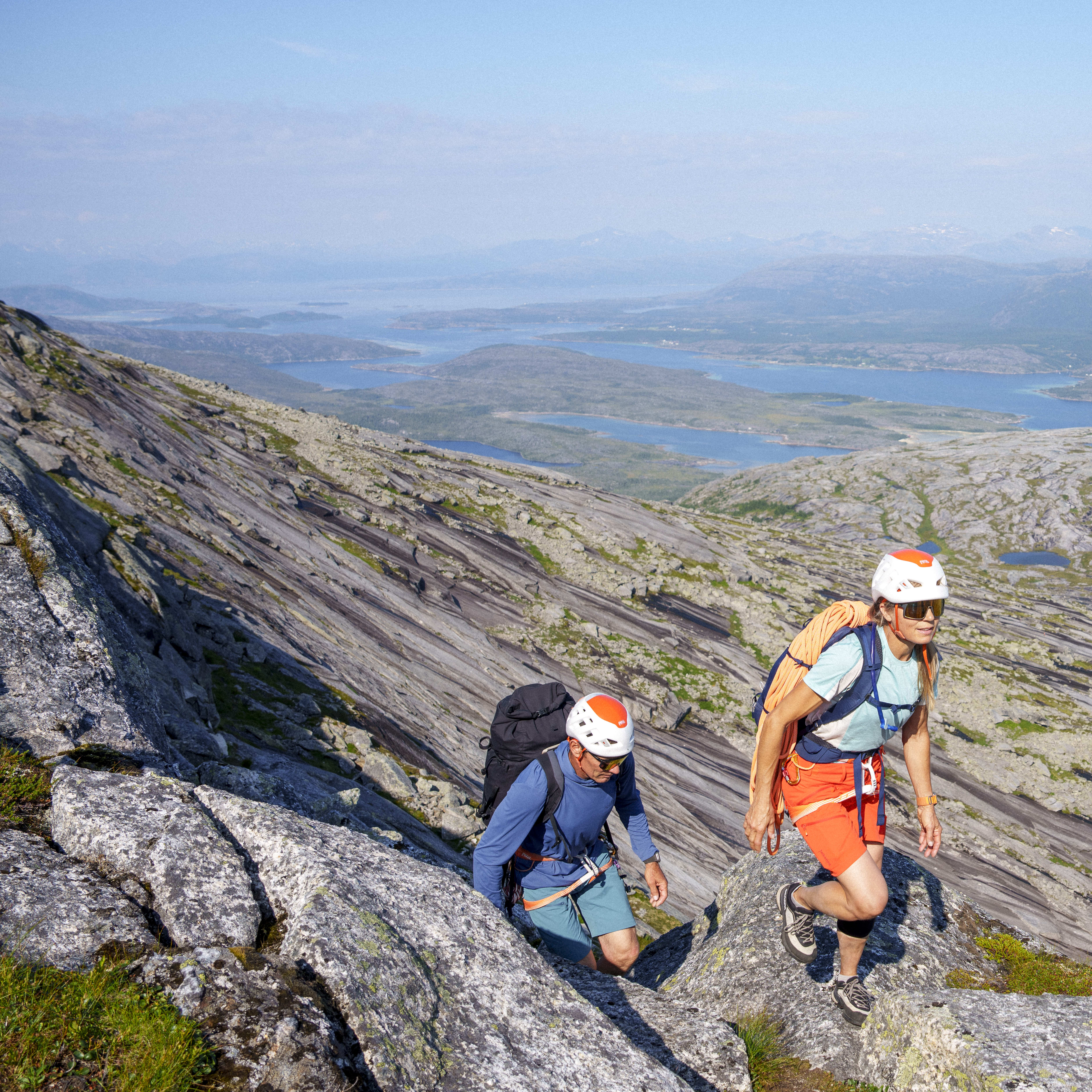 Two hikers climb a rocky mountain. One wears a large backpack, the other orange Norrøna shorts and climbing gear. Water and mountains visible in the background.
