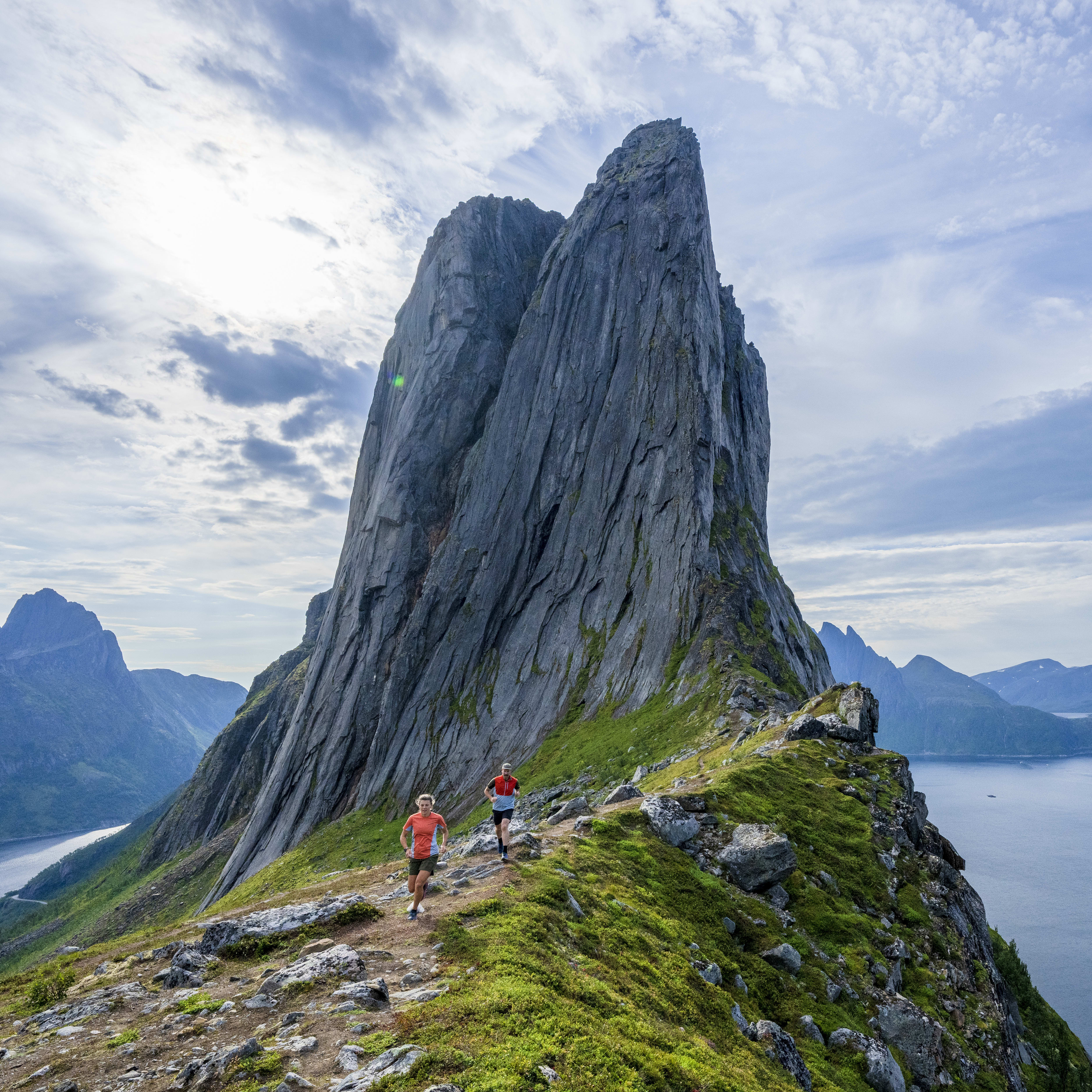 Two people in Norrøna gear running on a mountain path overlooking a fjord and other mountains. 
