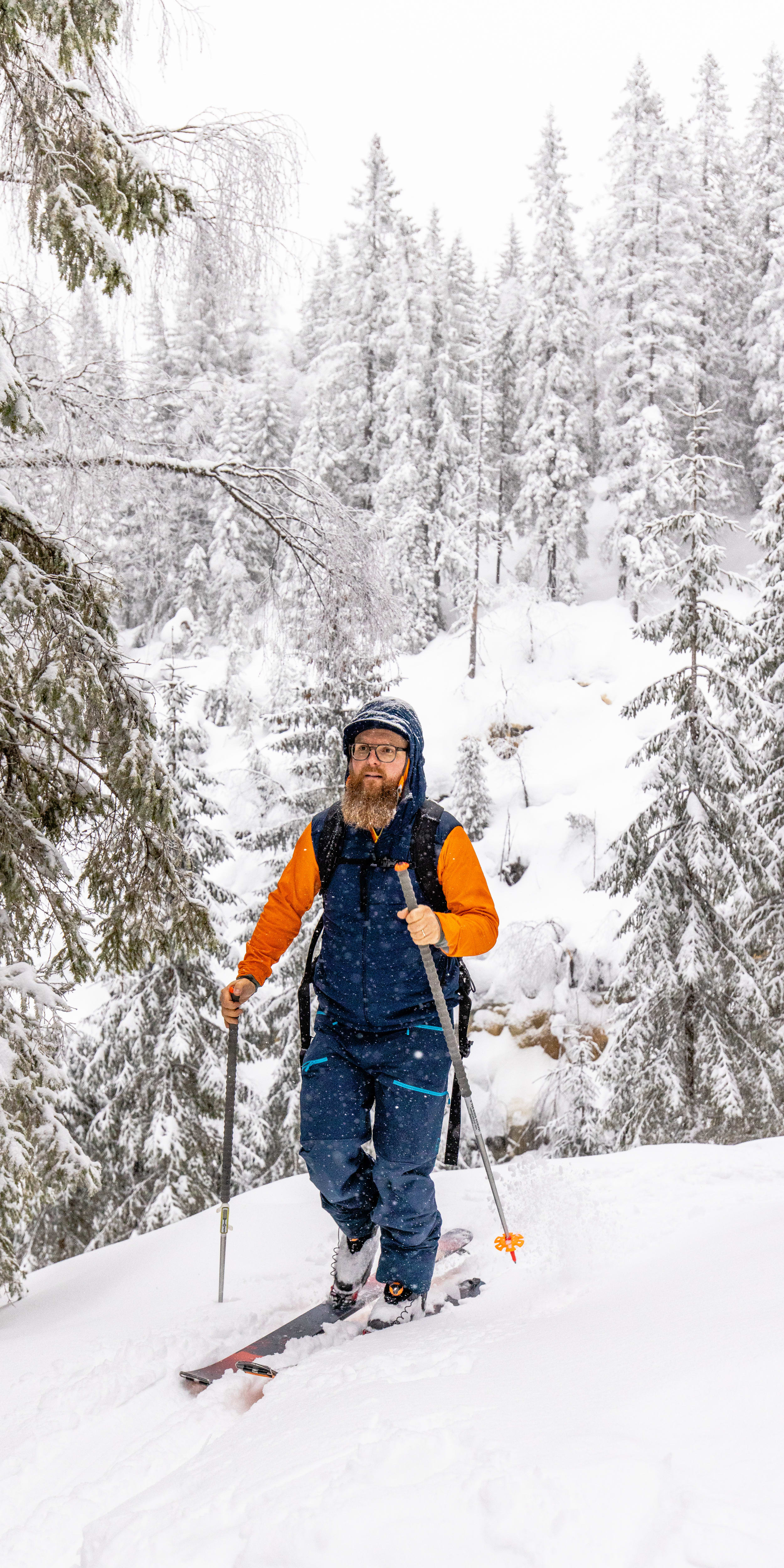 A man in Norrøna clothing backcountry skis through a snowy forest. 
