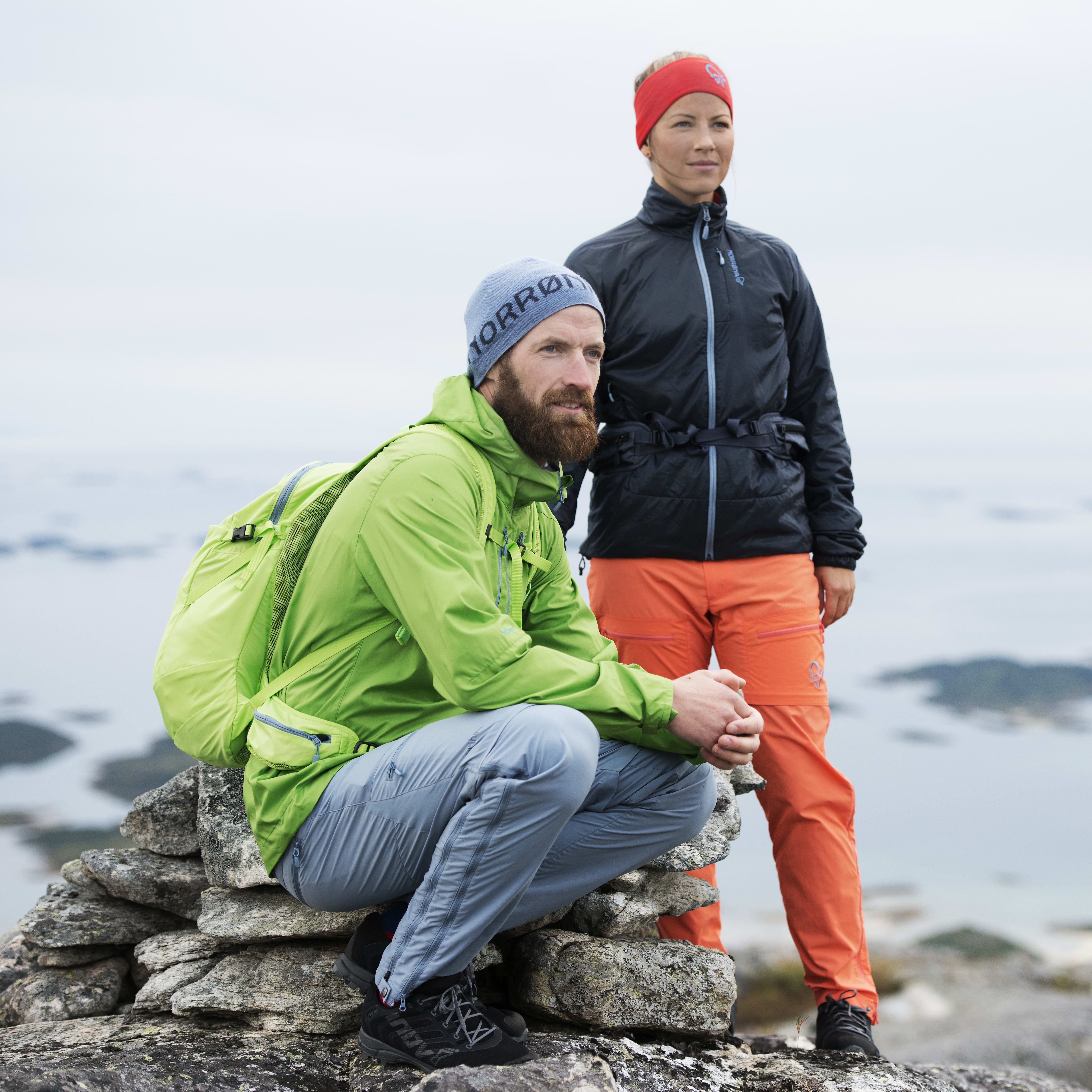 Two people wearing Norrøna clothing sit on a rocky mountaintop overlooking a body of water.  
