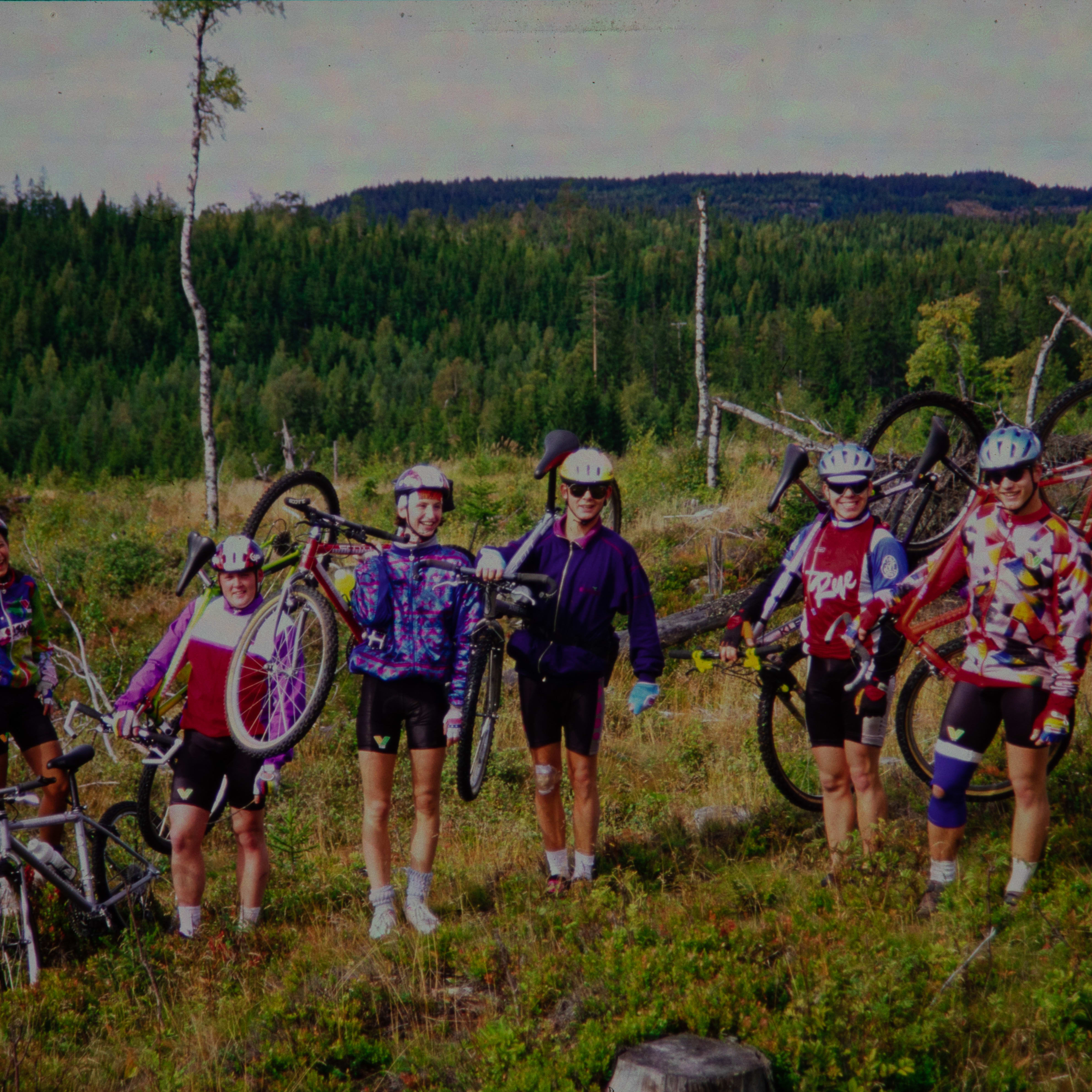 Five people in Norrøna clothing standing in a grassy field, holding and leaning on mountain bikes. A line of trees is in the background. There is a fallen log in the foreground.