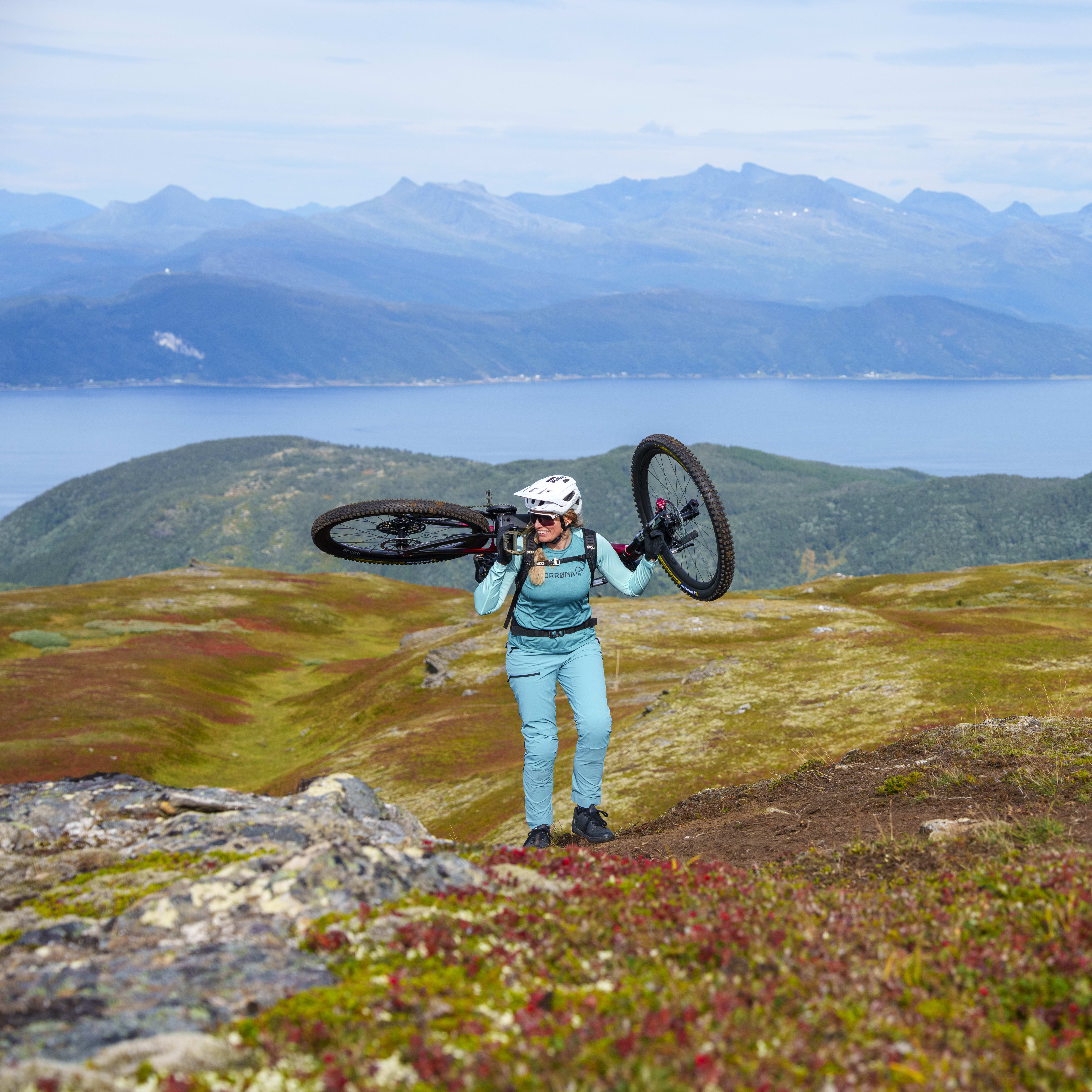 A woman in Norrøna mountain bike clothing carries a bicycle over her shoulders on a grassy hilltop, with mountains and a body of water in the background.
