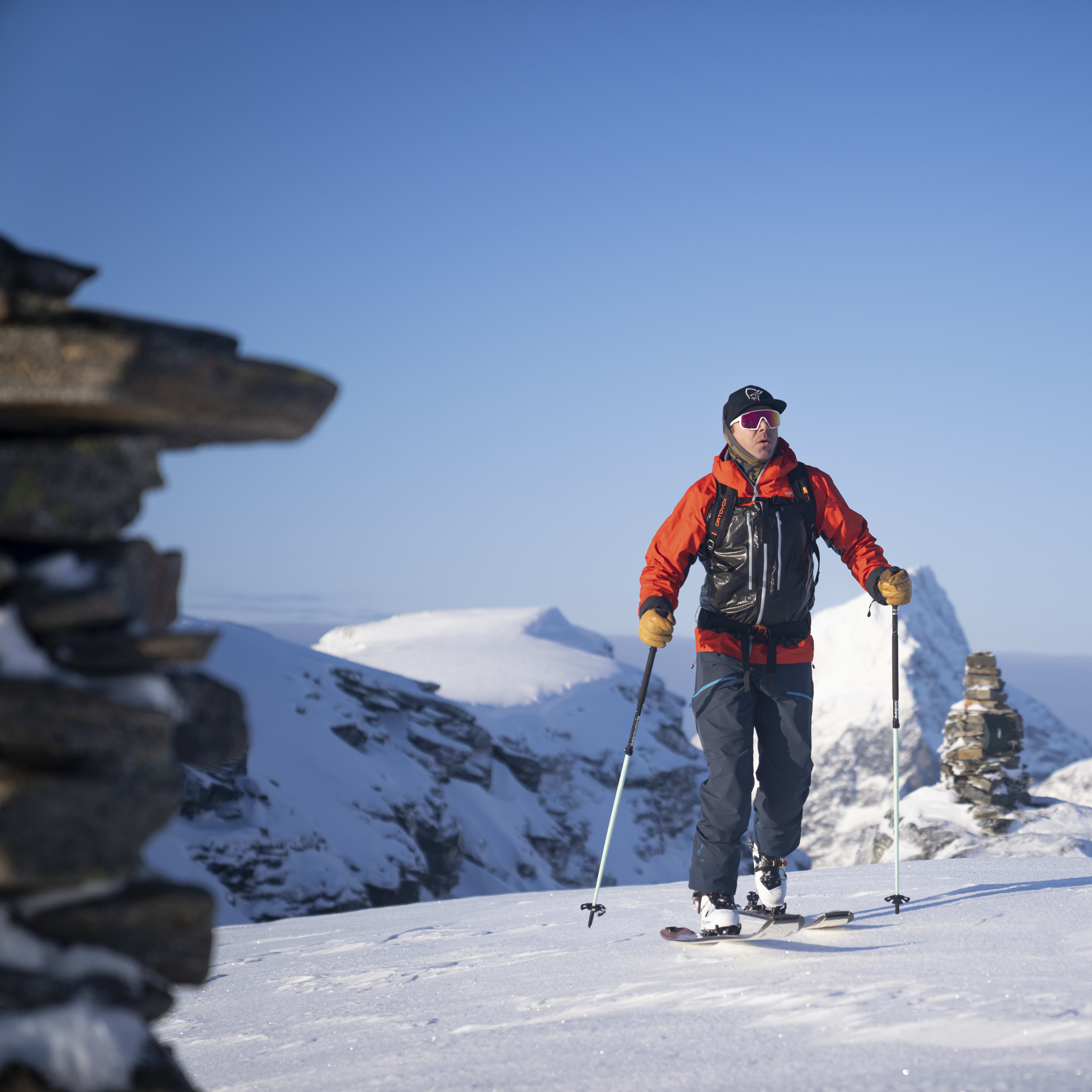 A skier in Norrøna gear (orange jacket, blue pants) skis on snow with mountain peaks in the background under a clear blue sky.