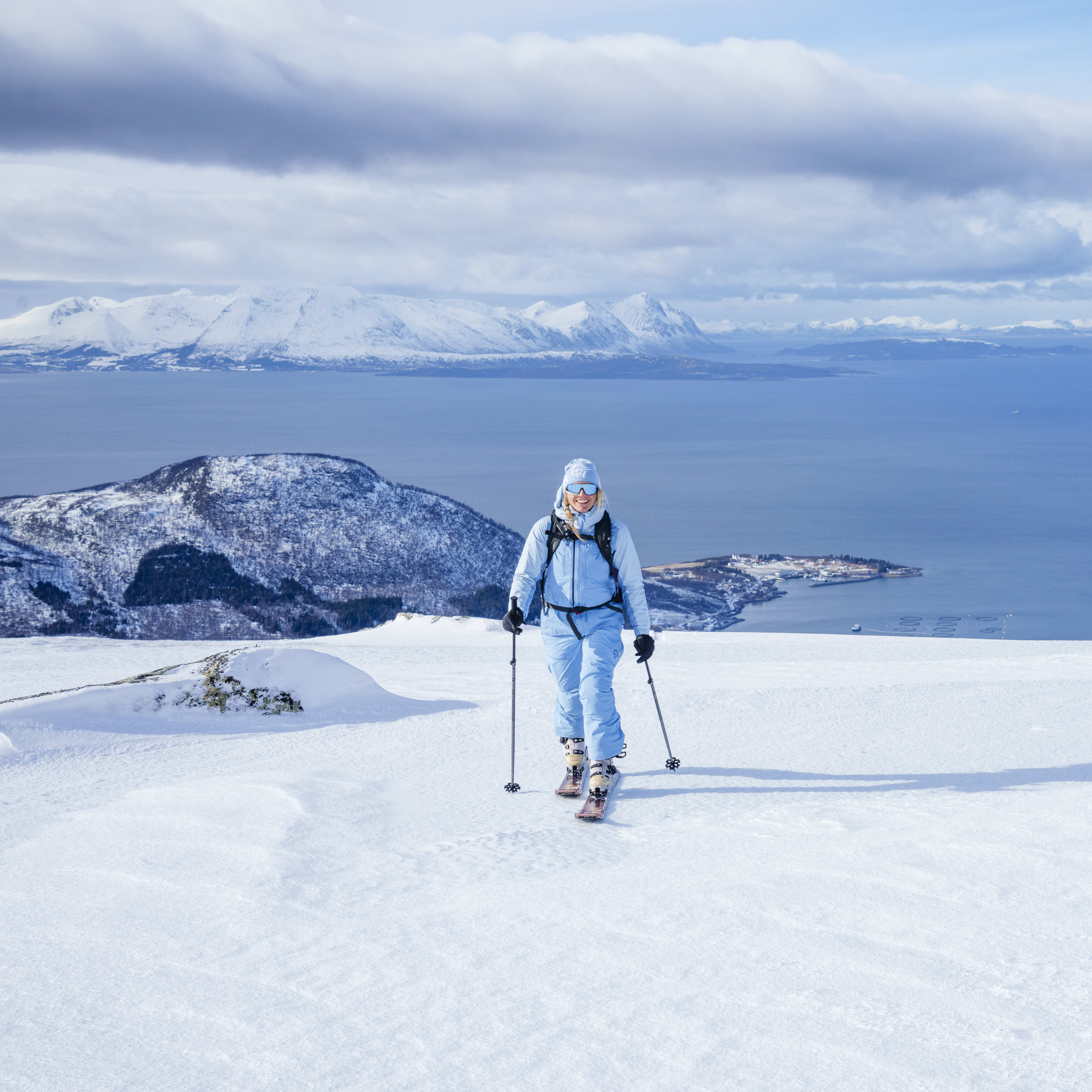 A skier in Norrøna blue outerwear stands on snow-covered ground with a view of the ocean and mountains in the background.