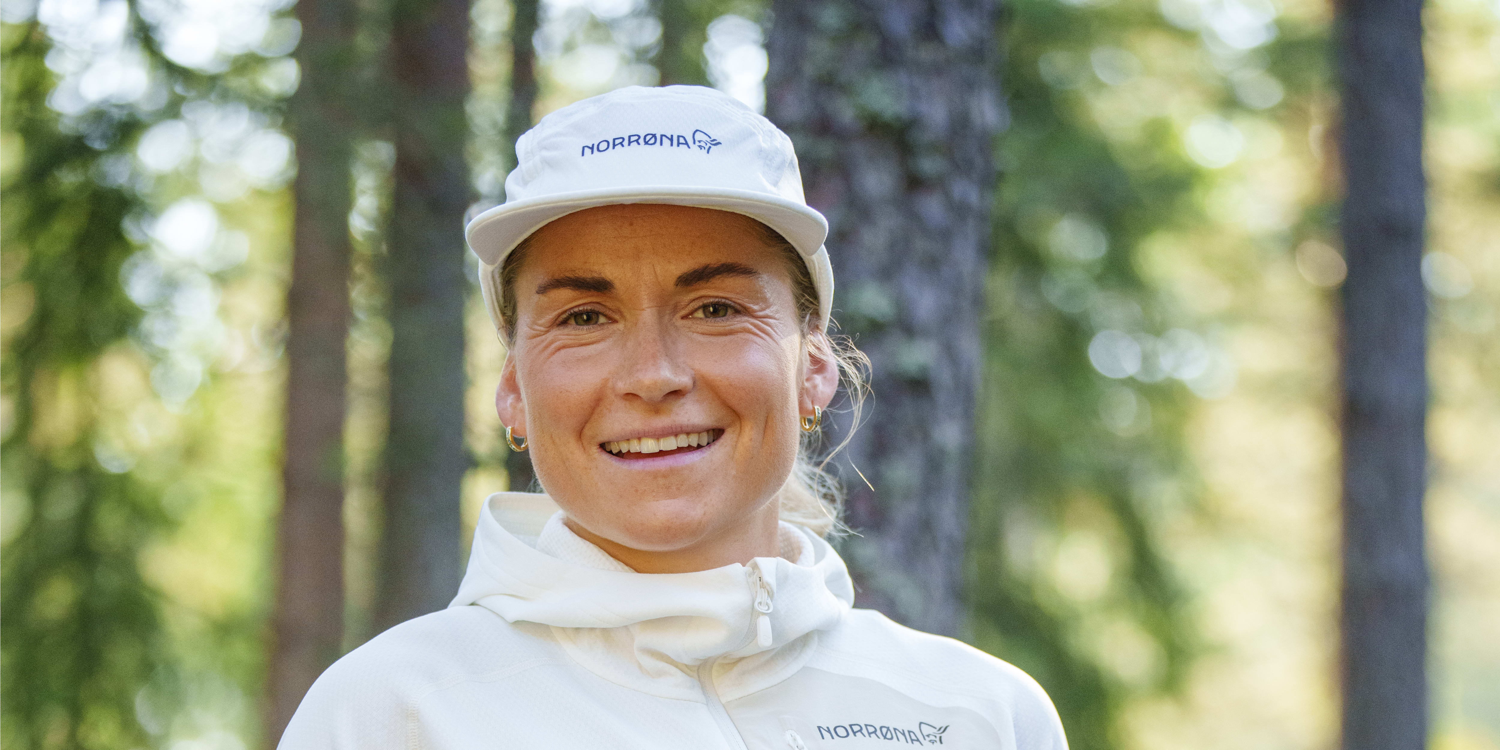 A smiling woman wearing a white Norrøna hoodie, a white Norrøna hat, and a green skirt, standing in a forest. 
