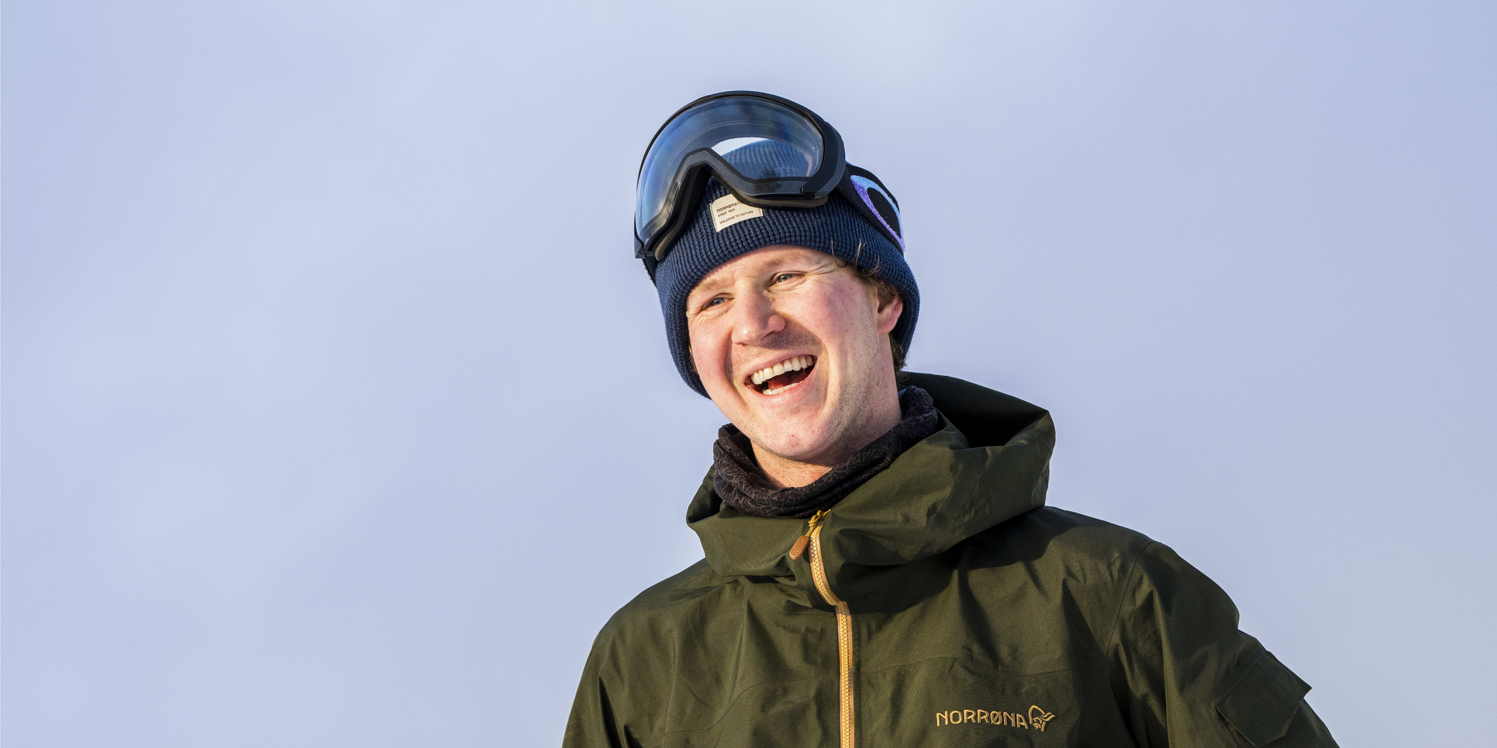 A smiling man wearing a Norrøna jacket and ski pants, holding ski poles in a snowy landscape.