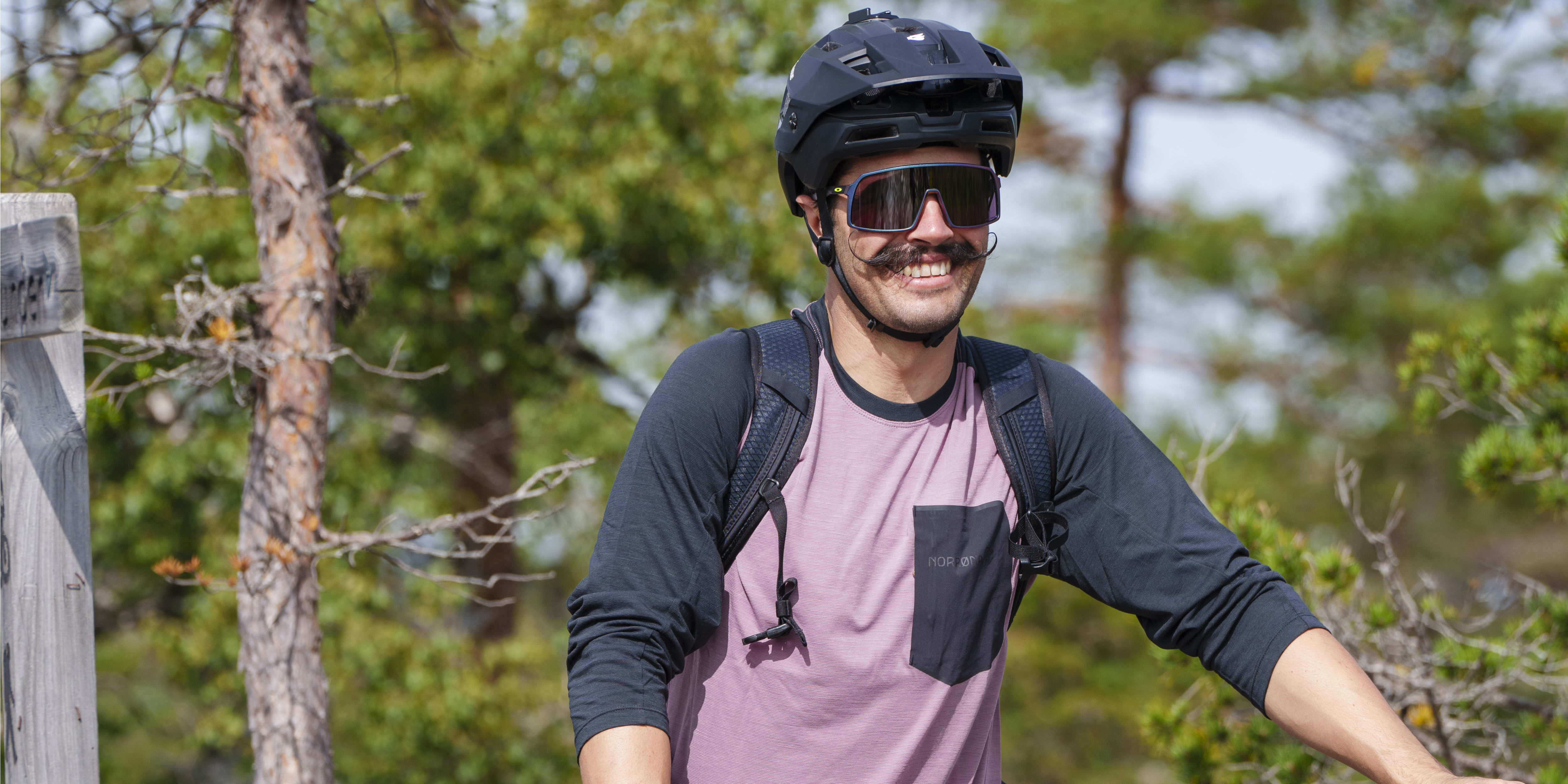 A person wearing a Norrøna shirt and pants is standing with a mountain bike in a forest. The bike is a Commençal and has a red Fox FloatX2 suspension. The person is wearing a black helmet, sunglasses, and black shoes. 
