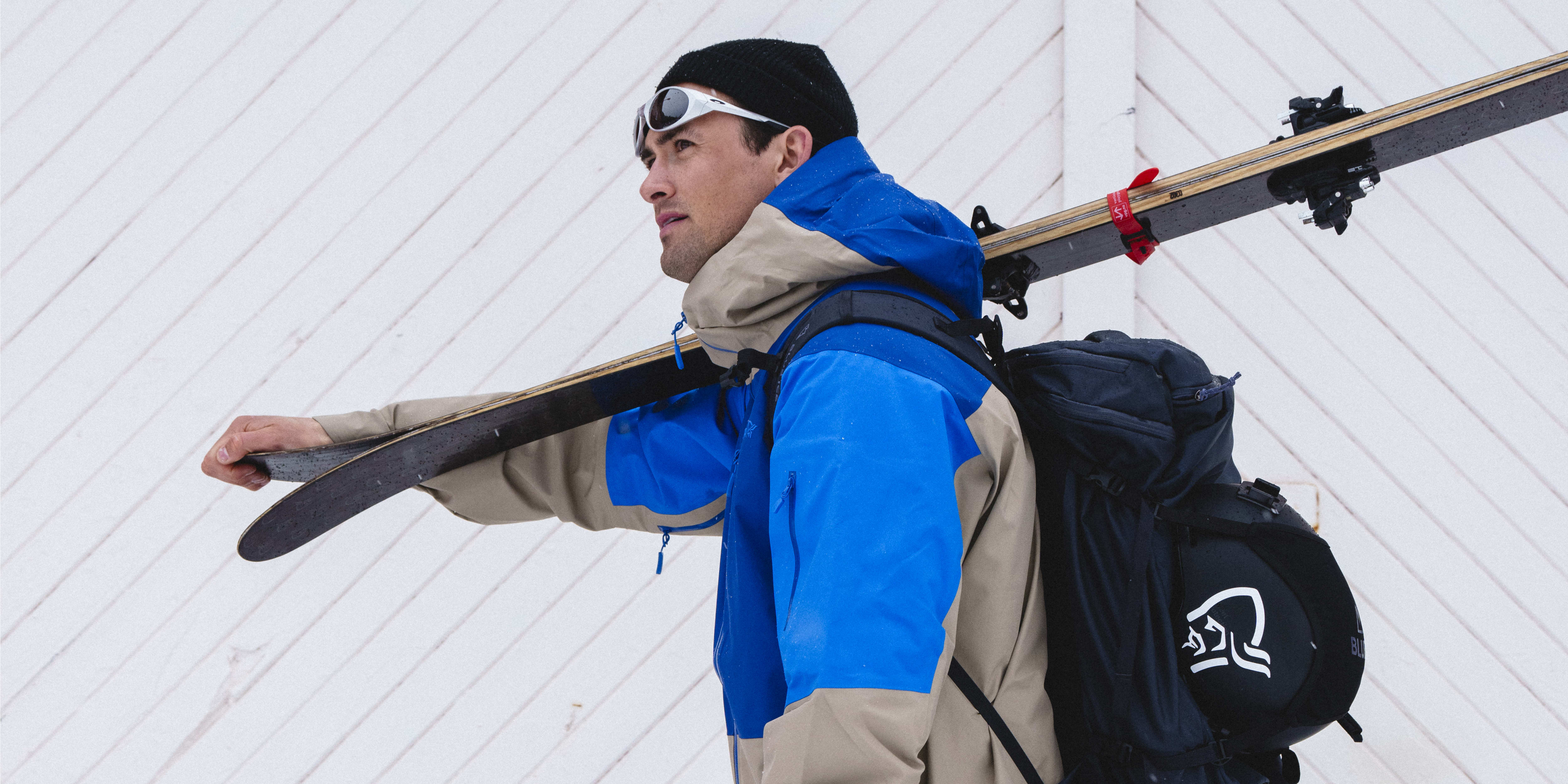 A man in a Norrøna ski jacket, pants, and boots, is walking in the snow while carrying skis and ski poles. He has a Norrøna backpack on. The man is wearing sunglasses and a black beanie. The image shows a wooden wall and some snow.