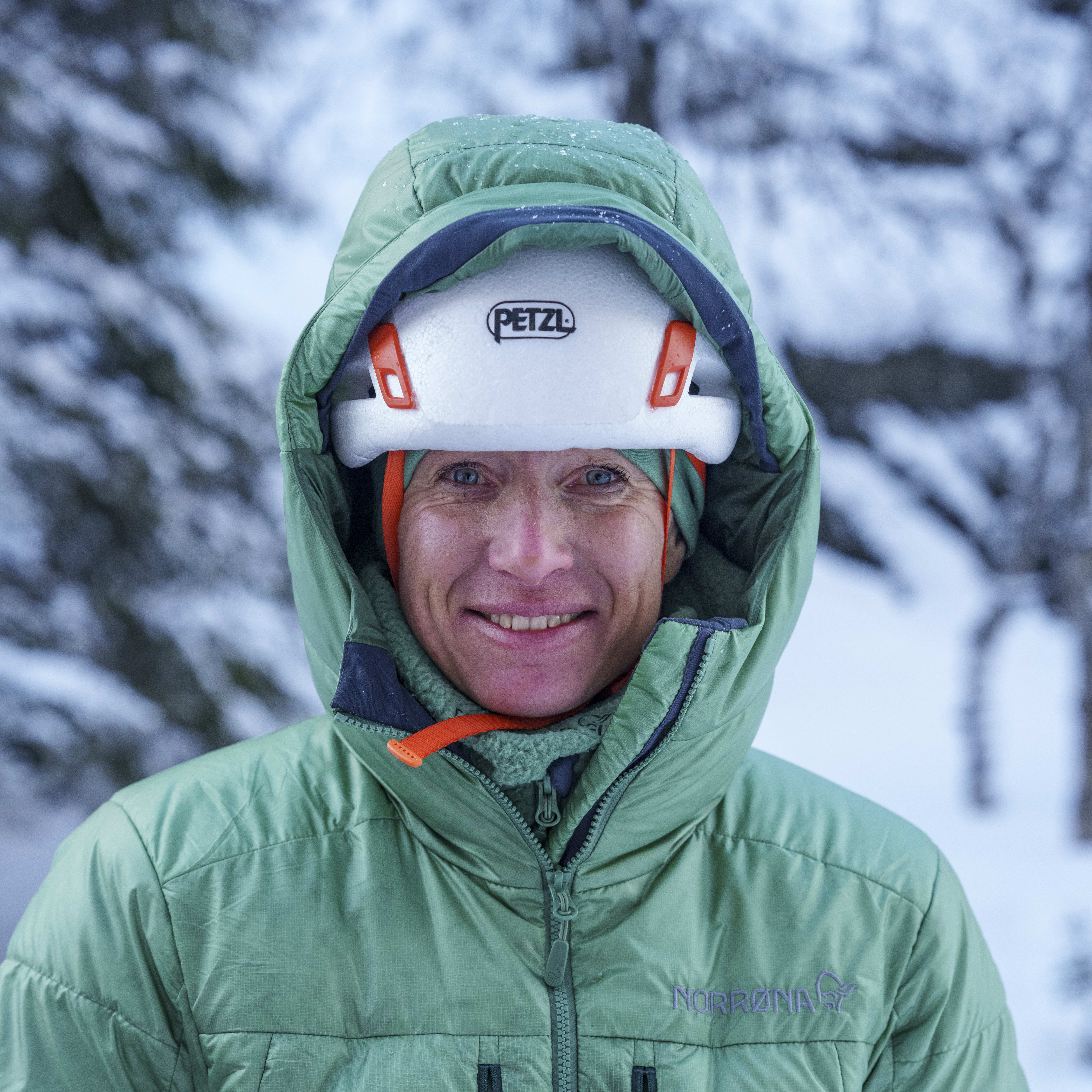 A person wearing a green Norrøna jacket and a white helmet with the word Petzl on it, smiles for the camera.