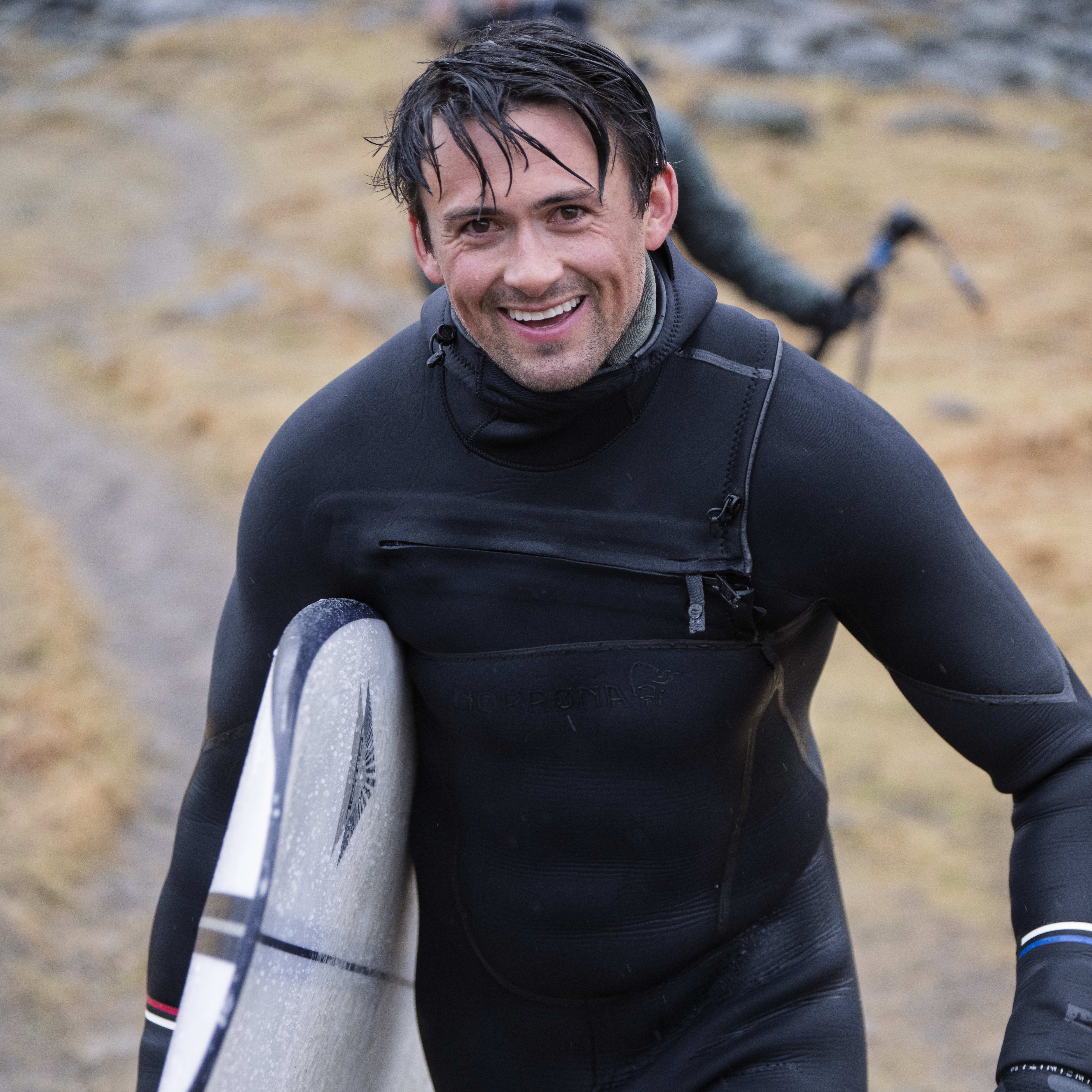 Man smiling, wearing a Norrøna wetsuit, holding a surfboard.