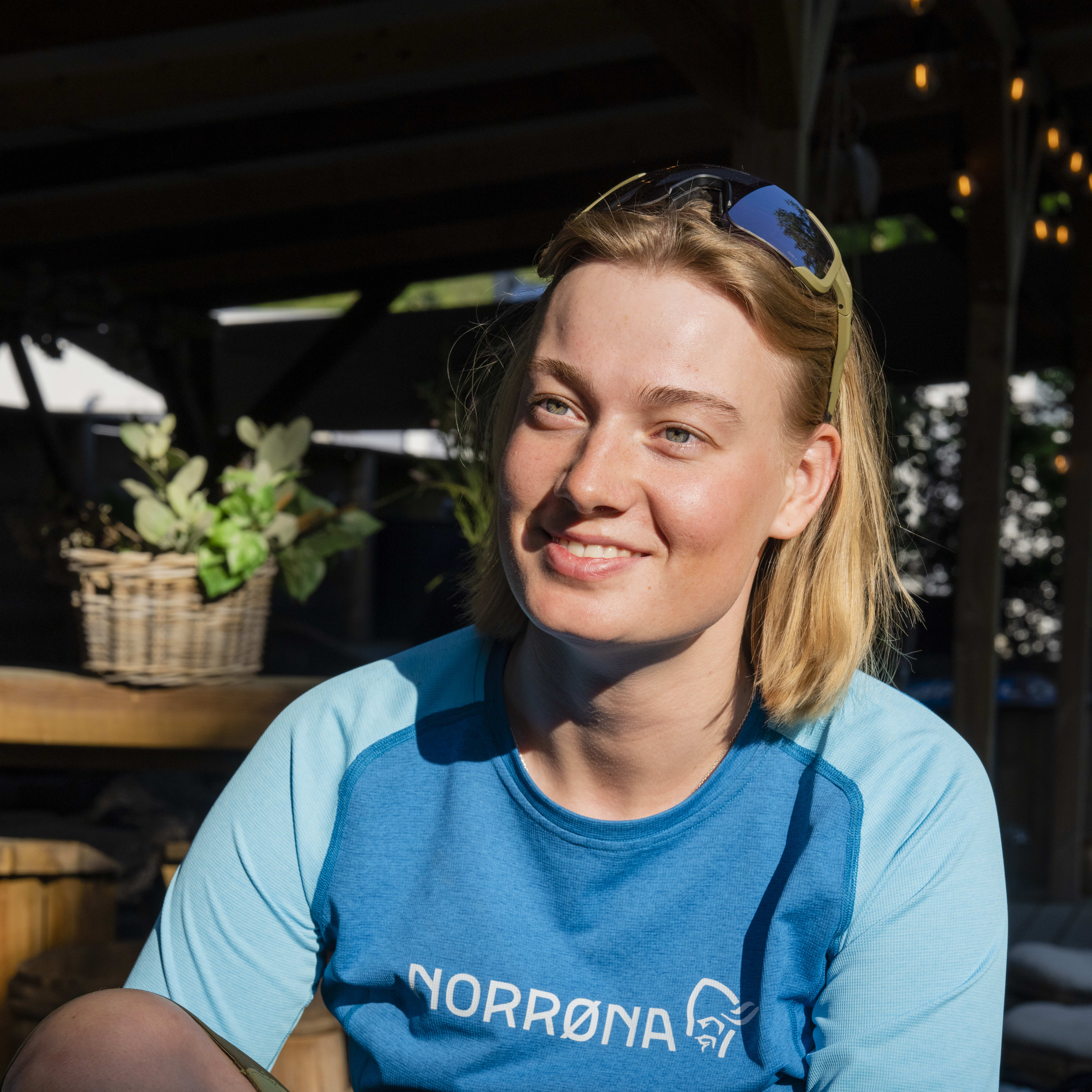 A woman wearing a Norrøna blue long-sleeved shirt and dark green pants, smiles at the camera. She has blonde hair and is wearing sunglasses. She is sitting with her legs bent and her hand resting on her thigh. The background is out of focus but appears to be wooden.