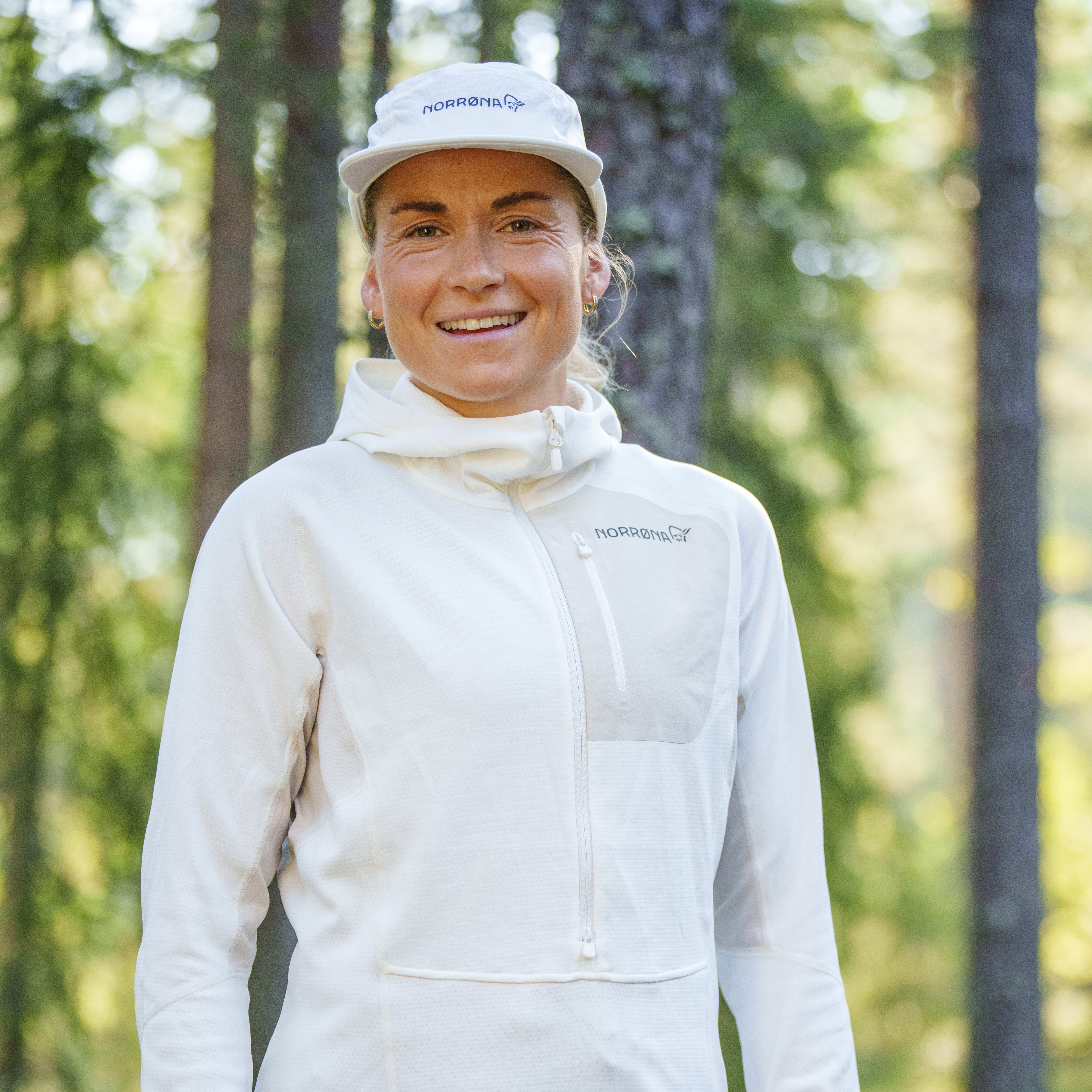 A smiling woman wearing a white Norrøna hoodie, a white Norrøna hat, and a green skirt, standing in a forest.