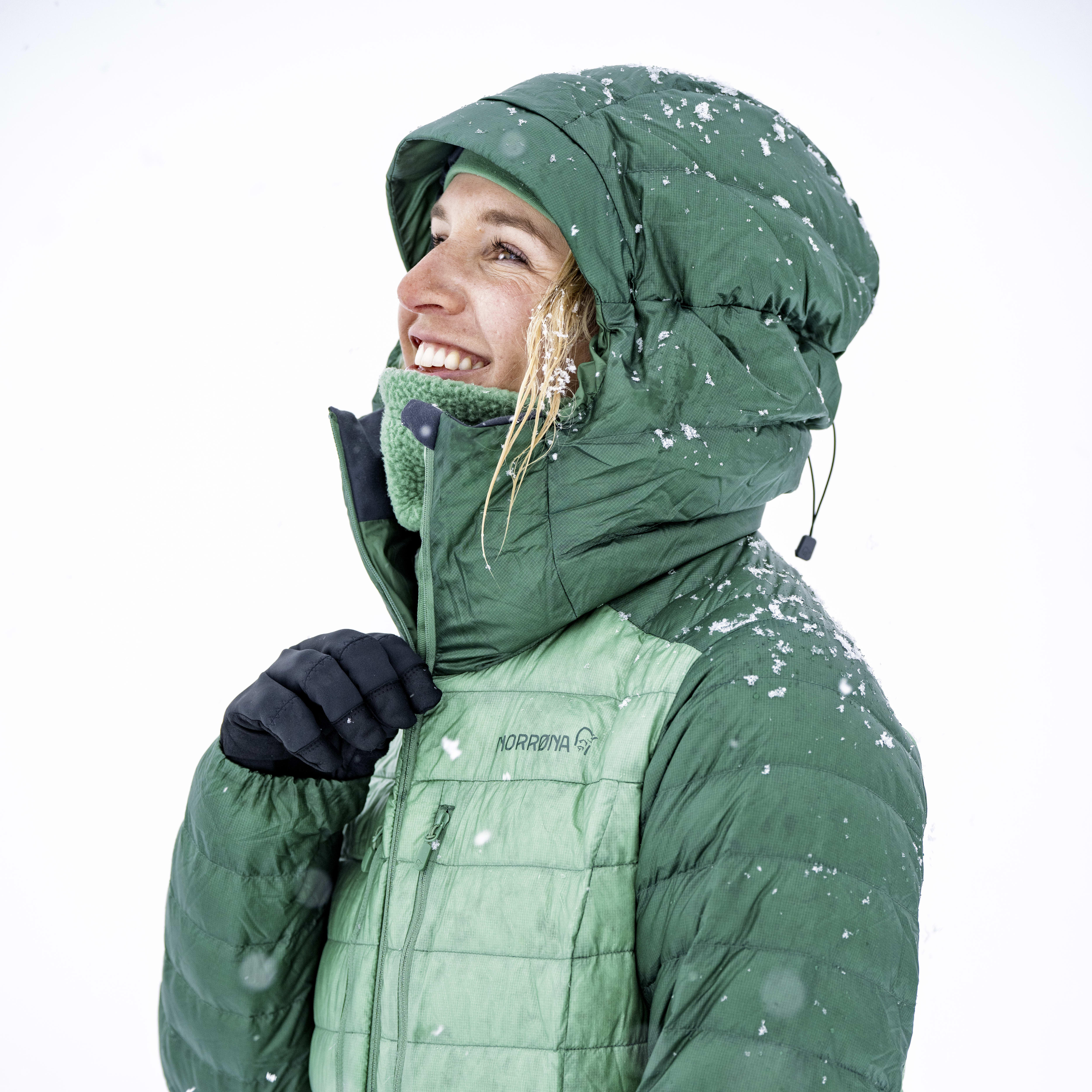 A woman wearing a green Norrøna jacket with a hood up, standing in snow.