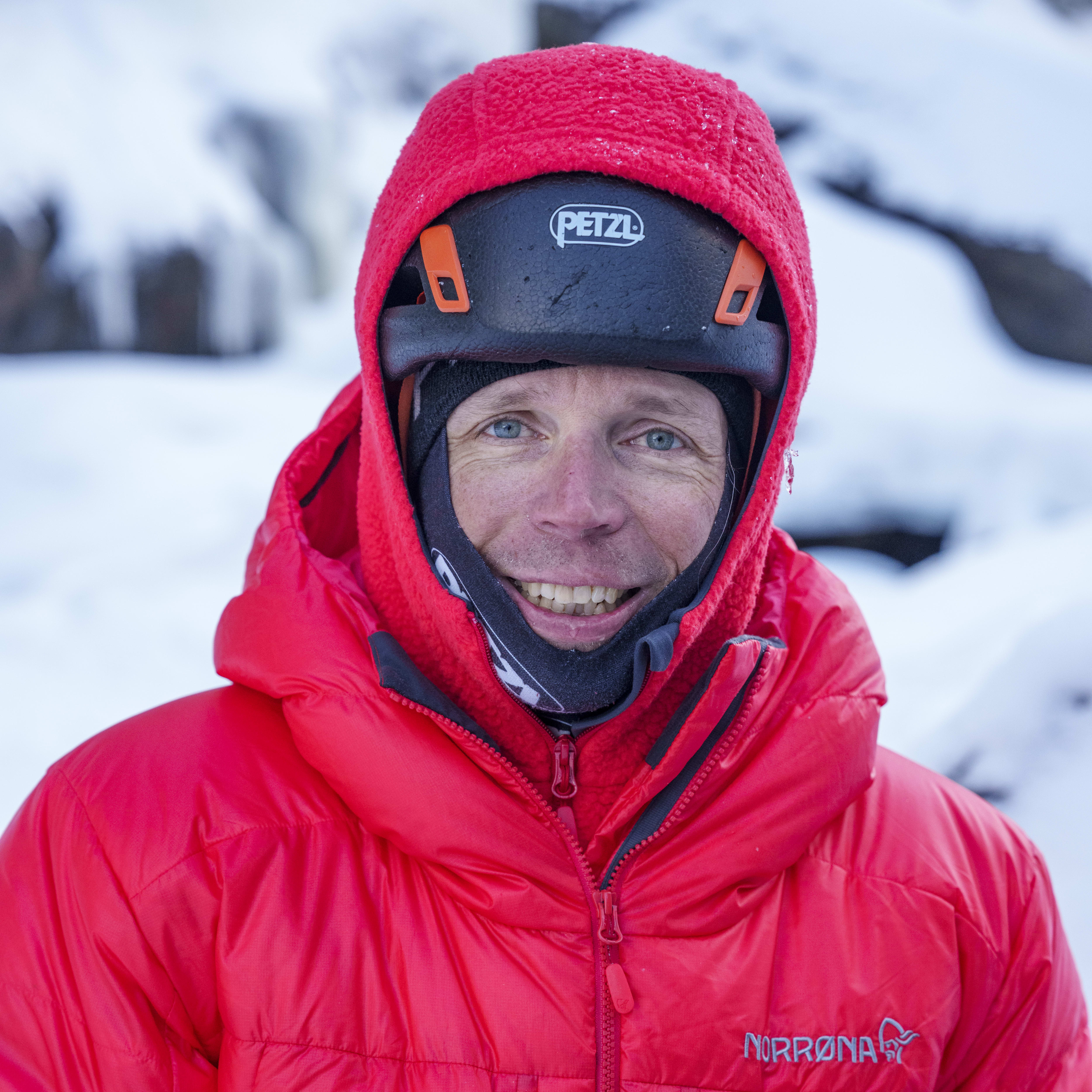 A man wearing a red Norrøna jacket and a black helmet smiles at the camera. He's holding a green water bottle. 
