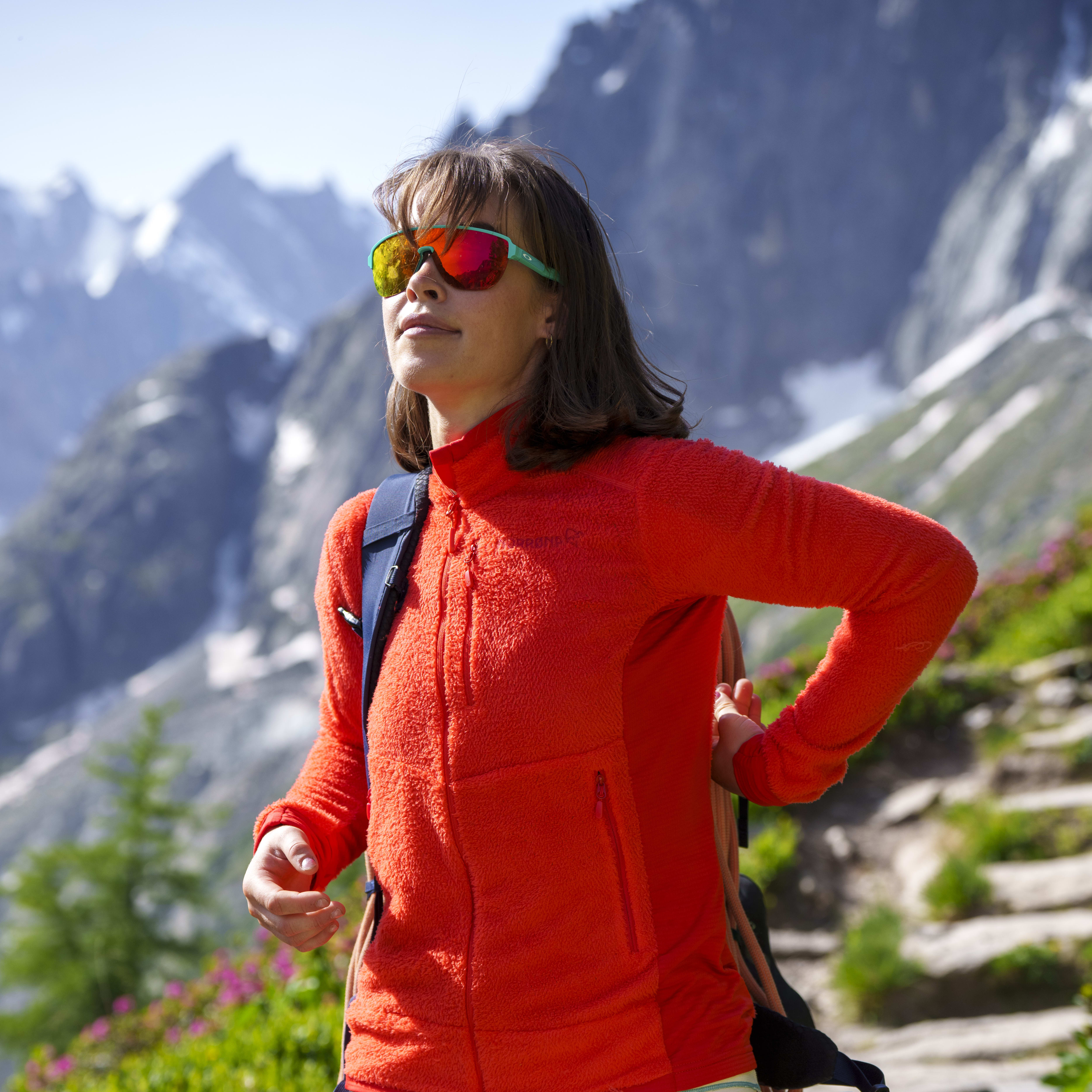 Woman hiking in the mountains wearing a bright orange Norrøna fleece and shorts, sunglasses, and a backpack.