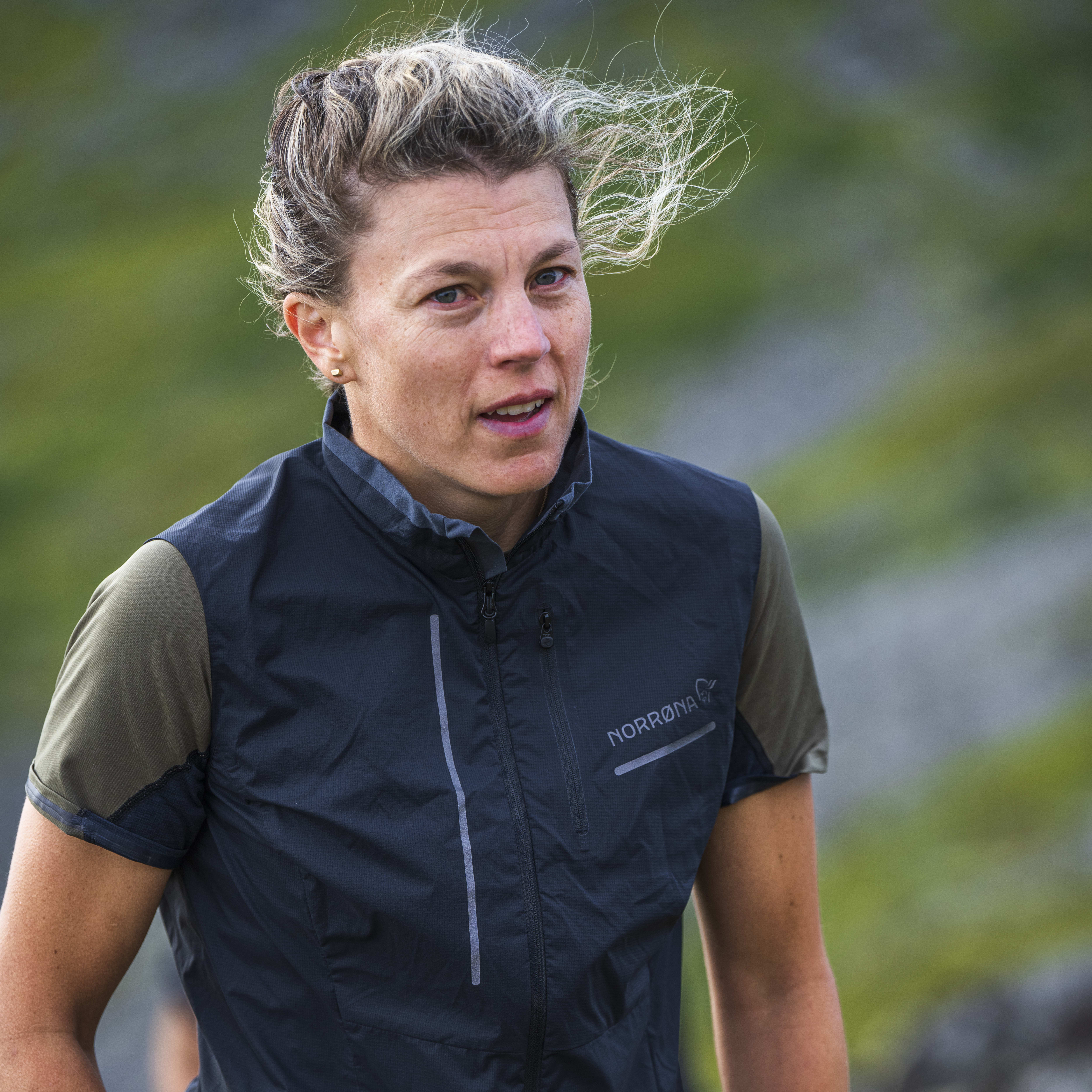A woman wearing a black Norrøna jacket and pants with a watch on her wrist.  She is standing in a field. 
