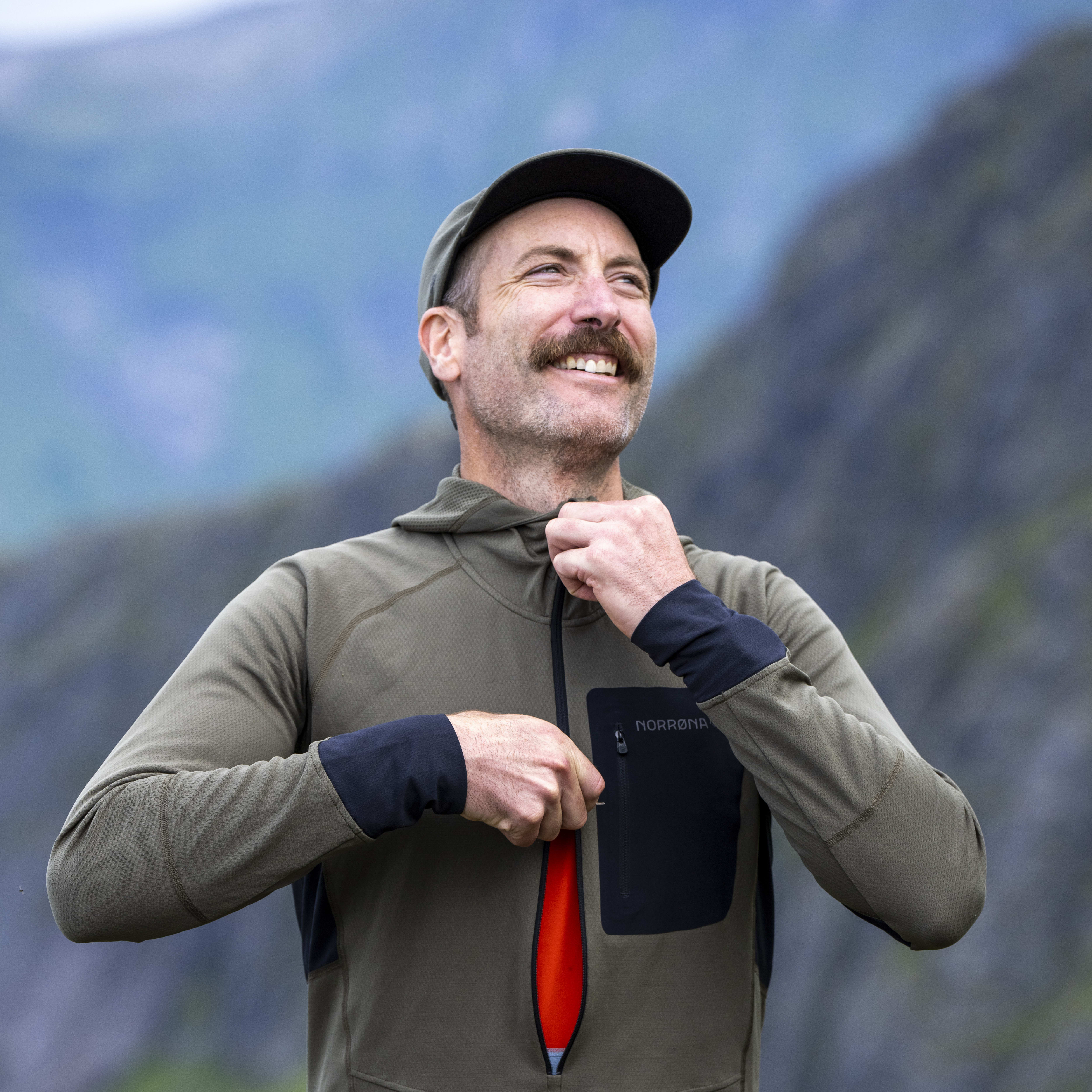 A man wearing a green Norrøna hoodie with a black zipper and a red pull tab, standing in front of a mountain landscape. 

