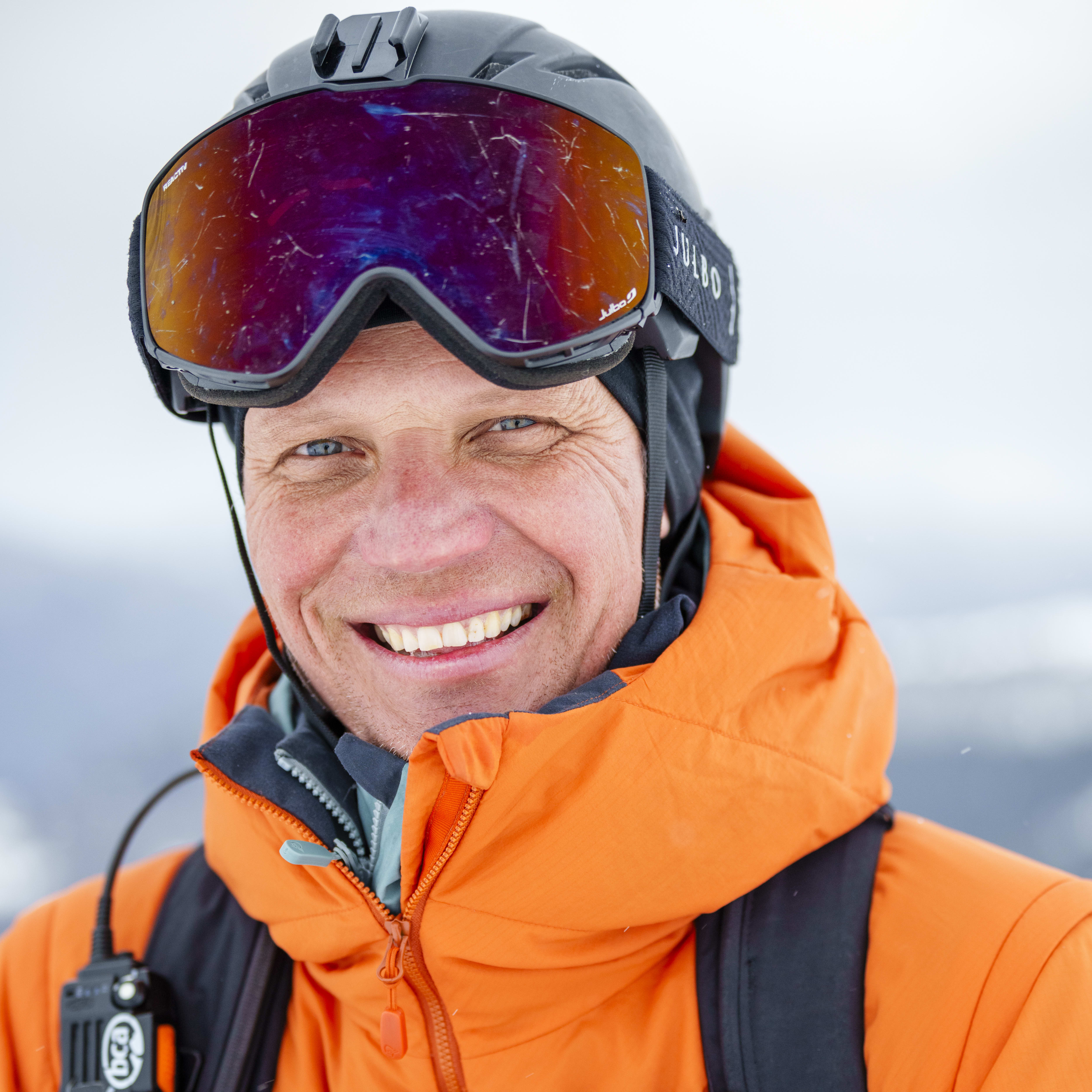 A smiling man wearing a helmet, goggles, and an orange Norrøna jacket is looking at the camera. A backpack and avalanche transceiver are visible.
