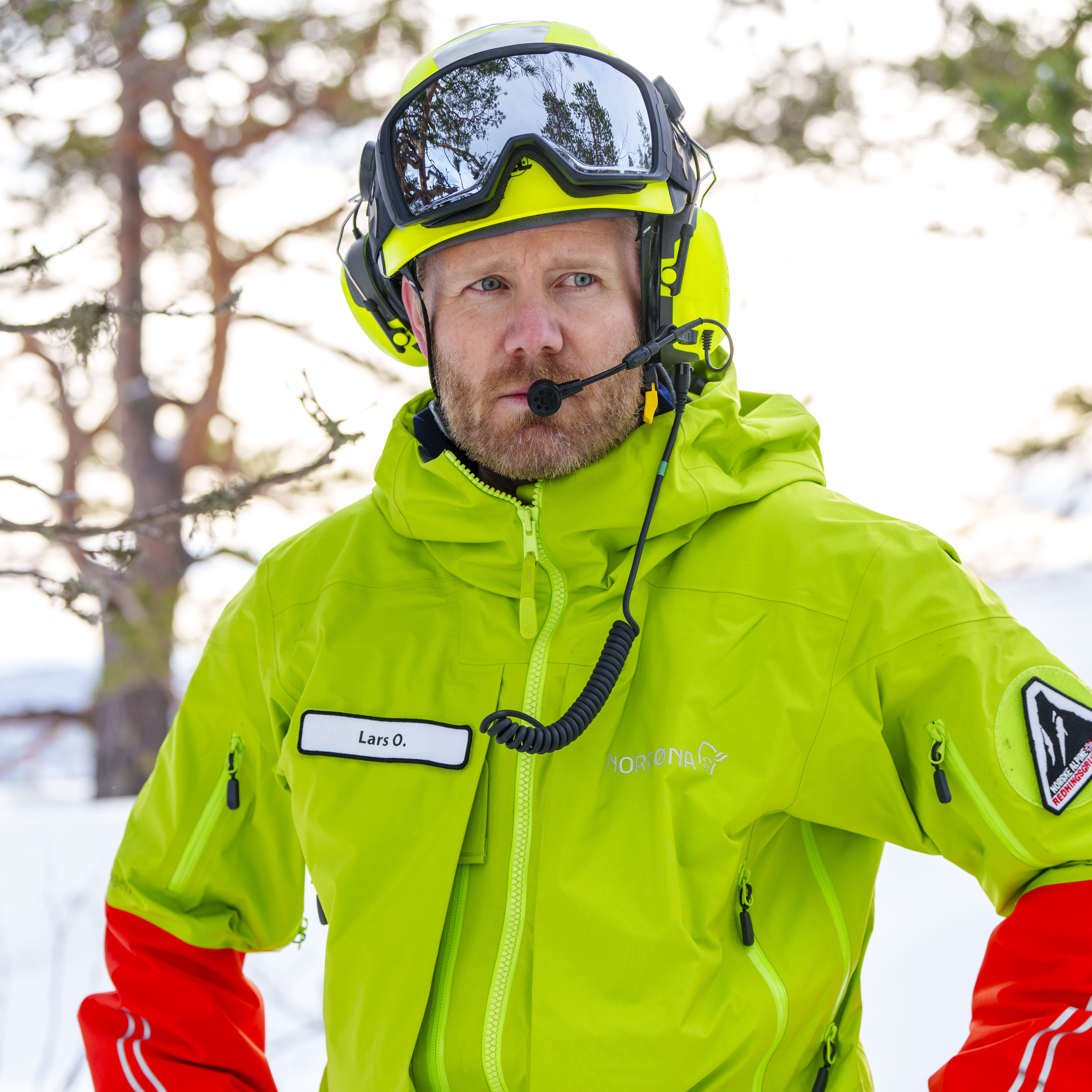 Man wearing a green Norrøna jacket, black gloves and a helmet with a headset, standing in a snowy outdoor environment. 
