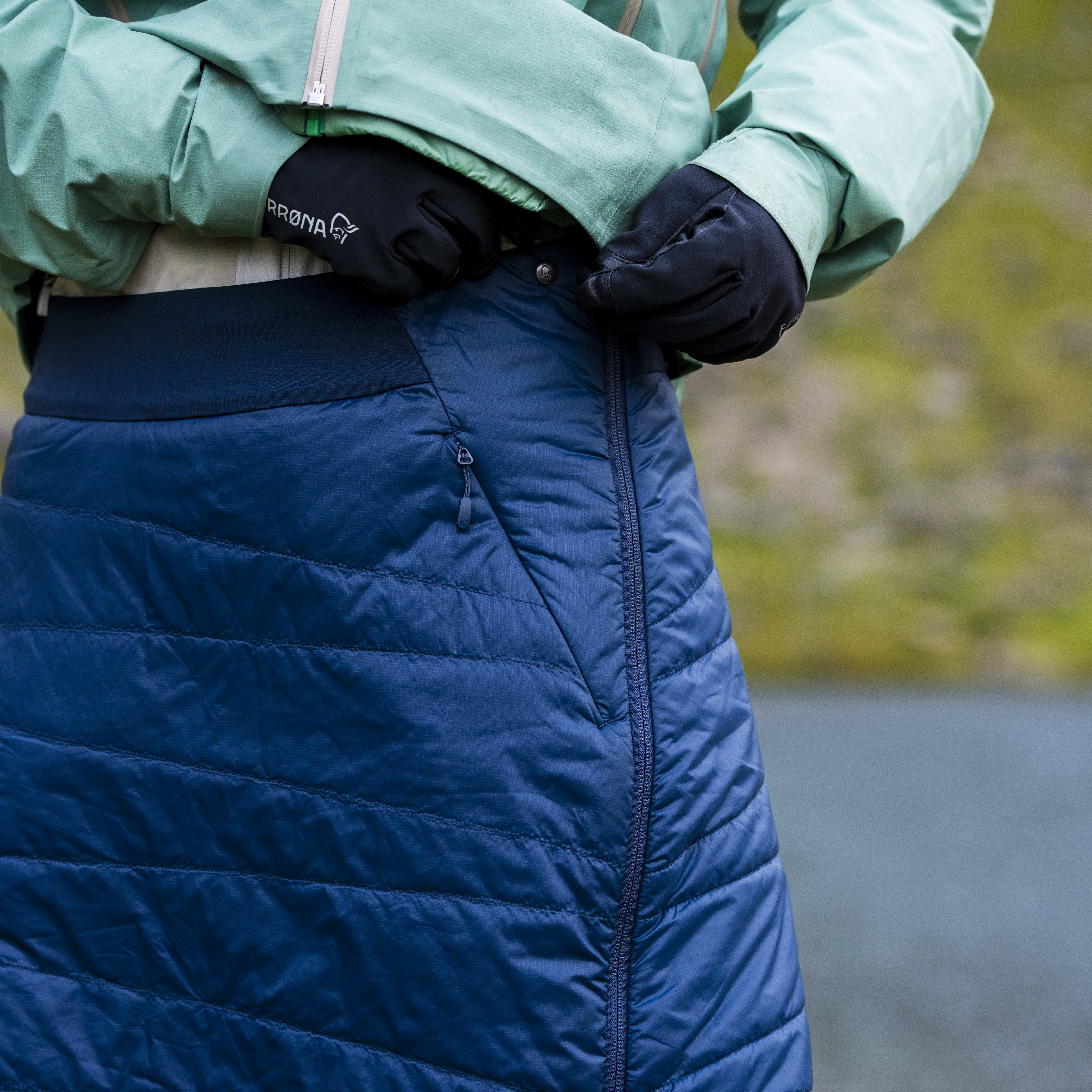 A woman is wearing a blue Norrøna insulated skirt. The skirt has a zipper on the side and a white logo.