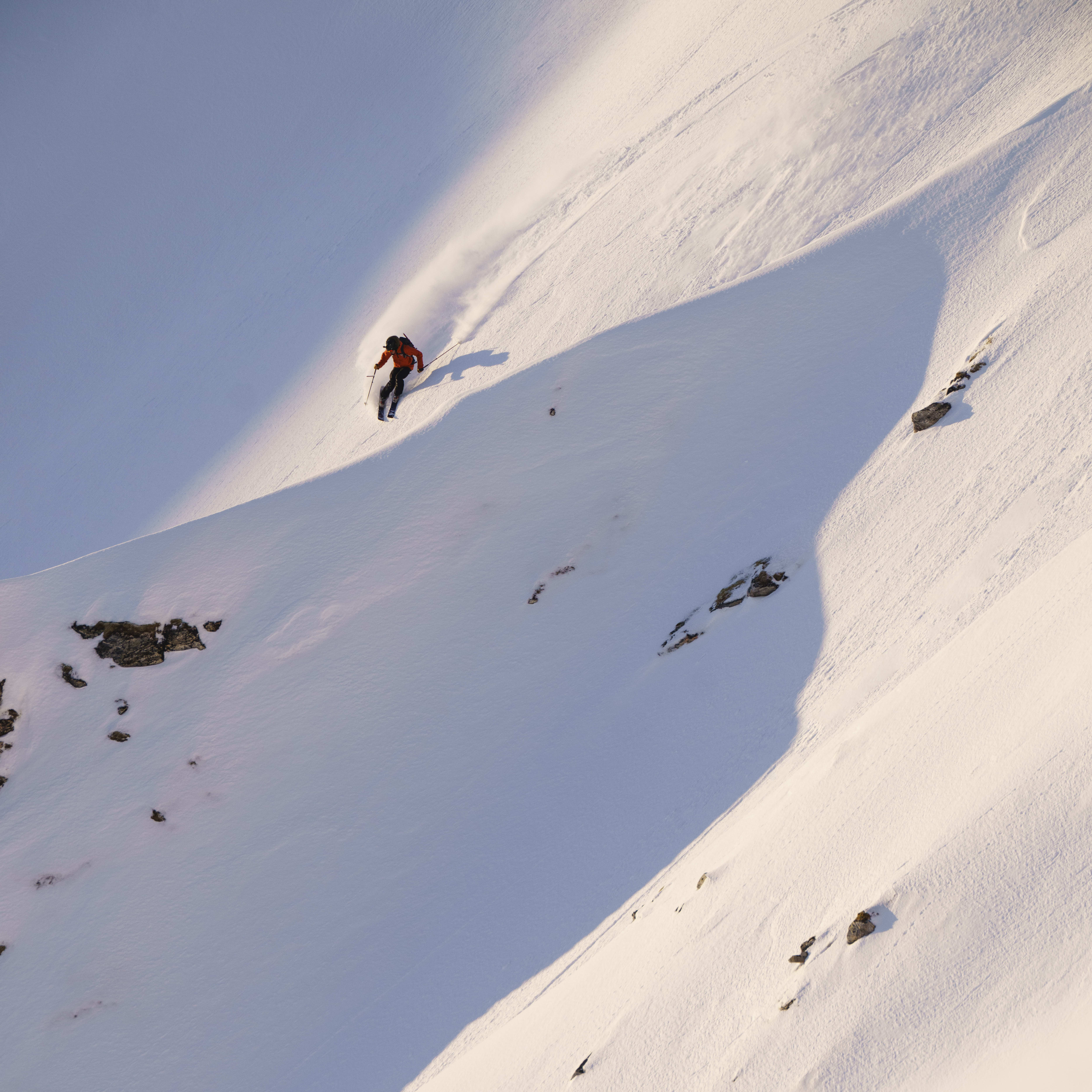 A skier descends a snow-covered mountain, wearing a red Norrøna jacket, carving tracks in the pristine powder on a sunny day.