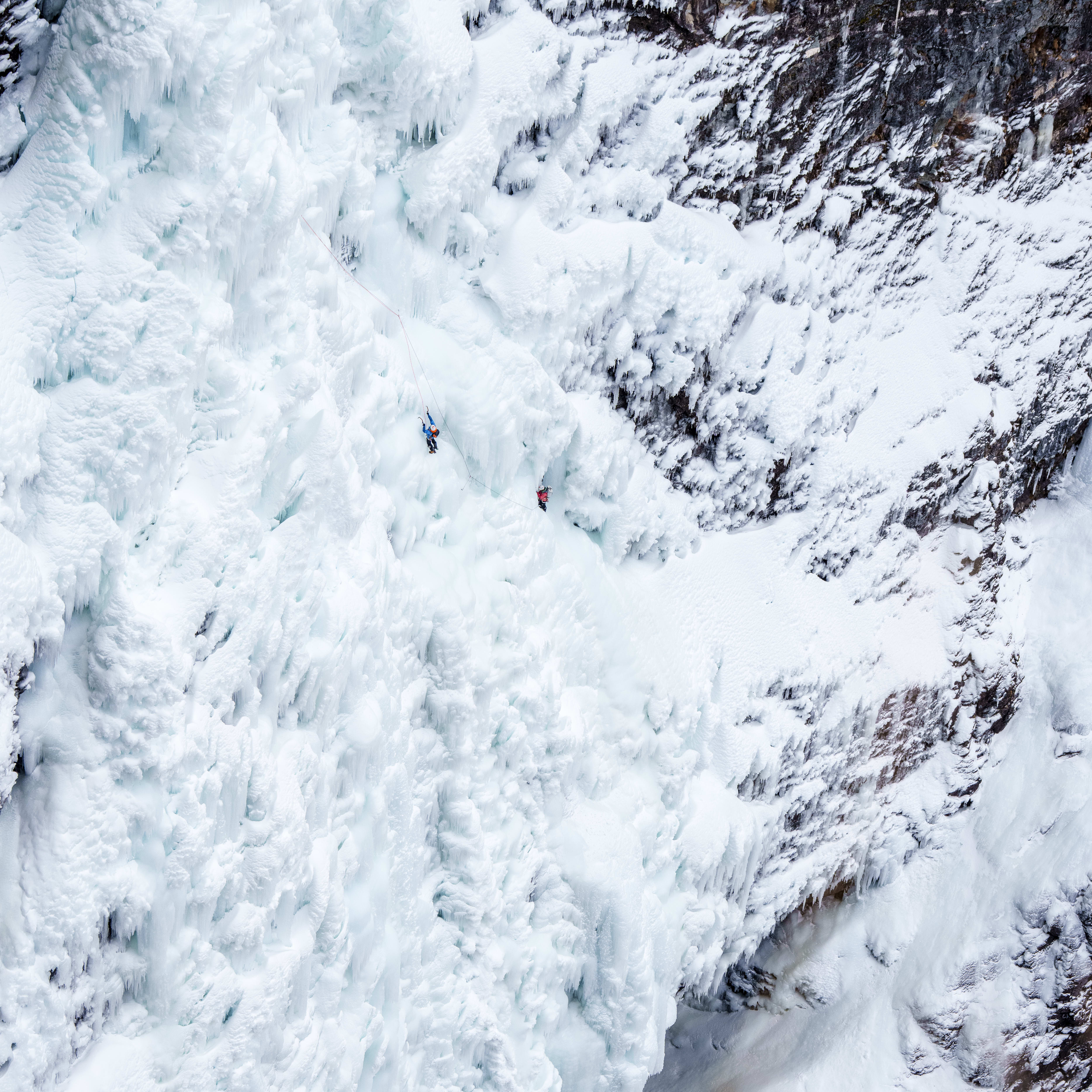 Two ice climbers ascend a steep, frozen waterfall. One wears a blue Norrøna jacket, the other, red.