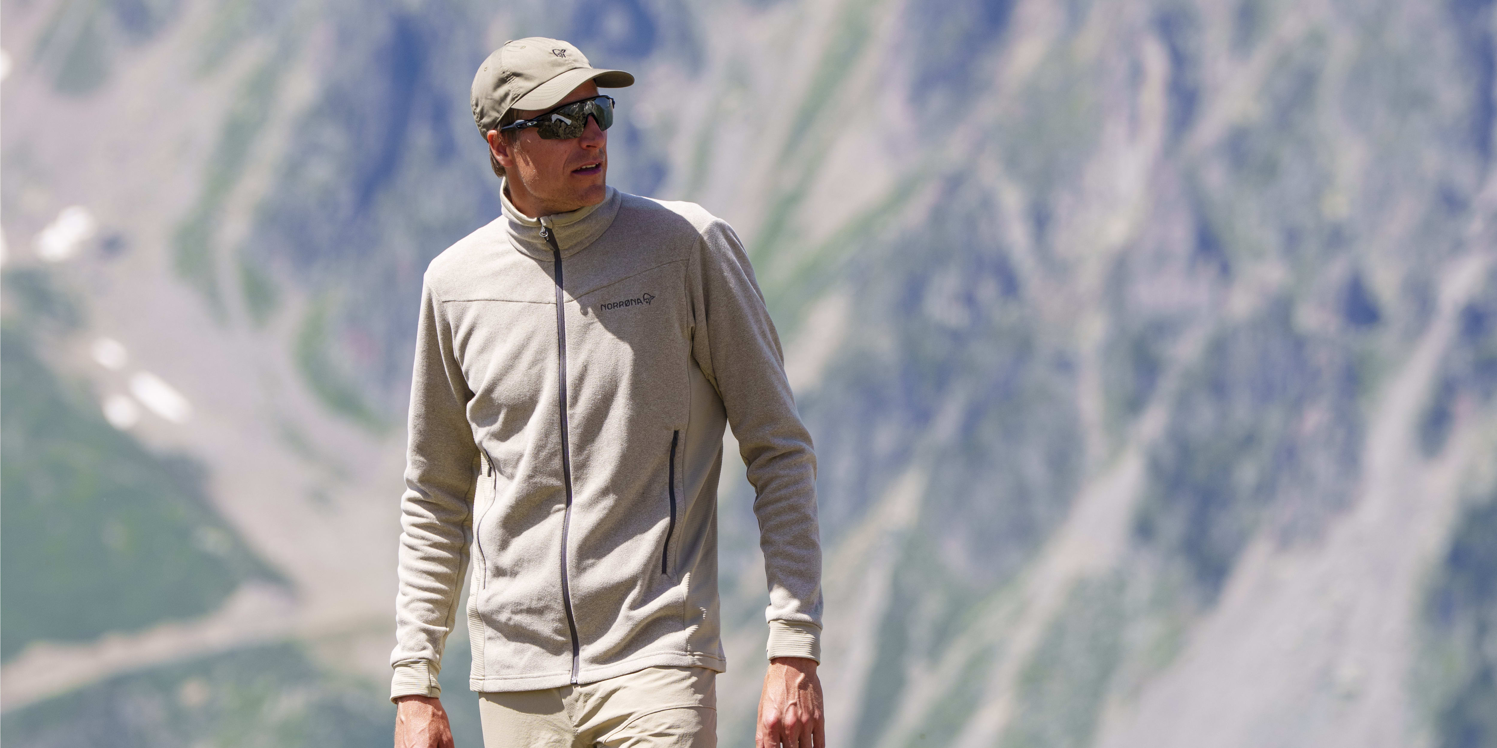 A man stands on a rocky peak wearing a Norrøna fleece and shorts. He wears sunglasses and a hat, with mountains in the background.
