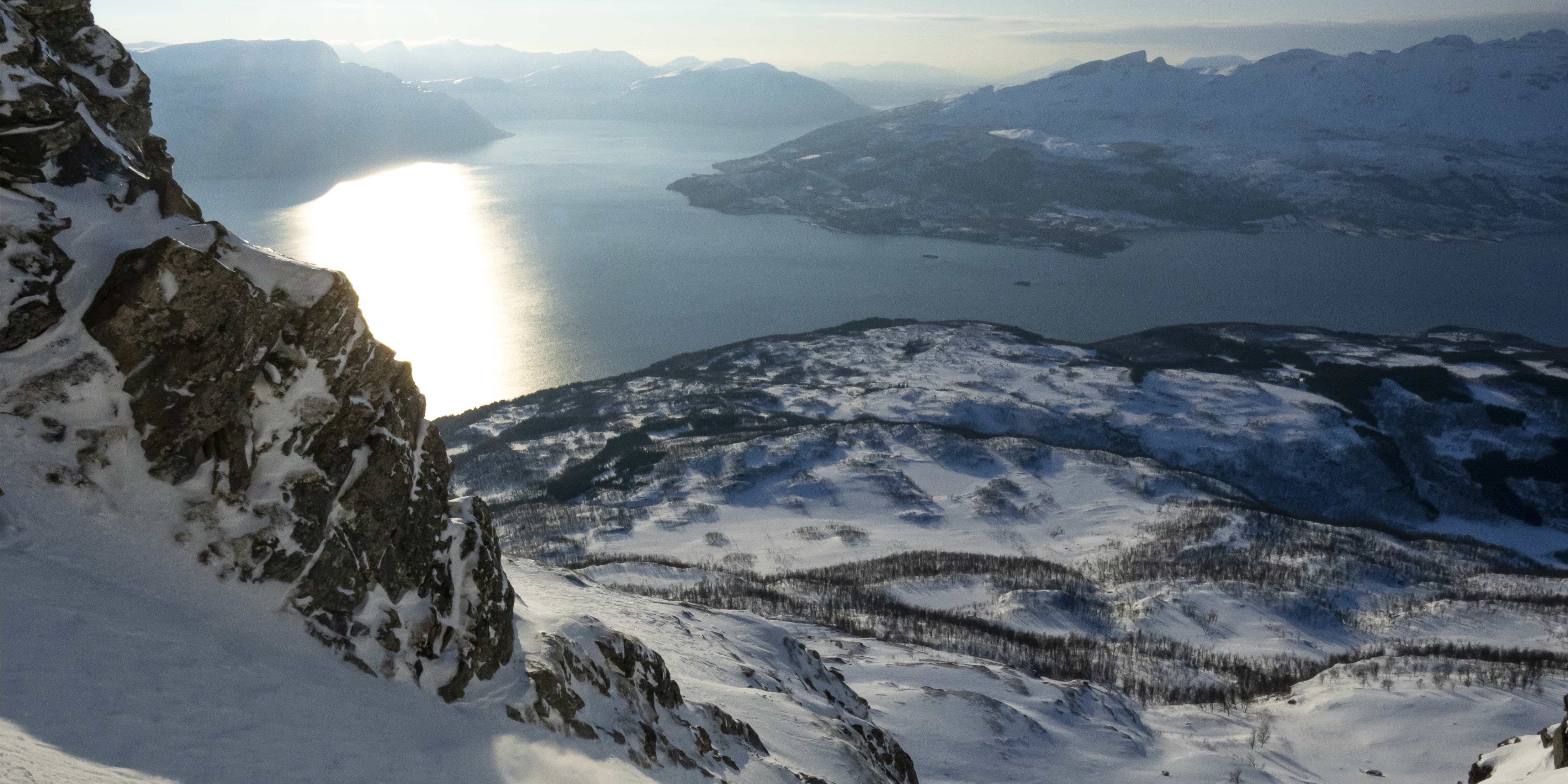 A skier in a red Norrøna jacket skis down a snowy mountain slope on a sunny day with a fjord and mountains in the background.