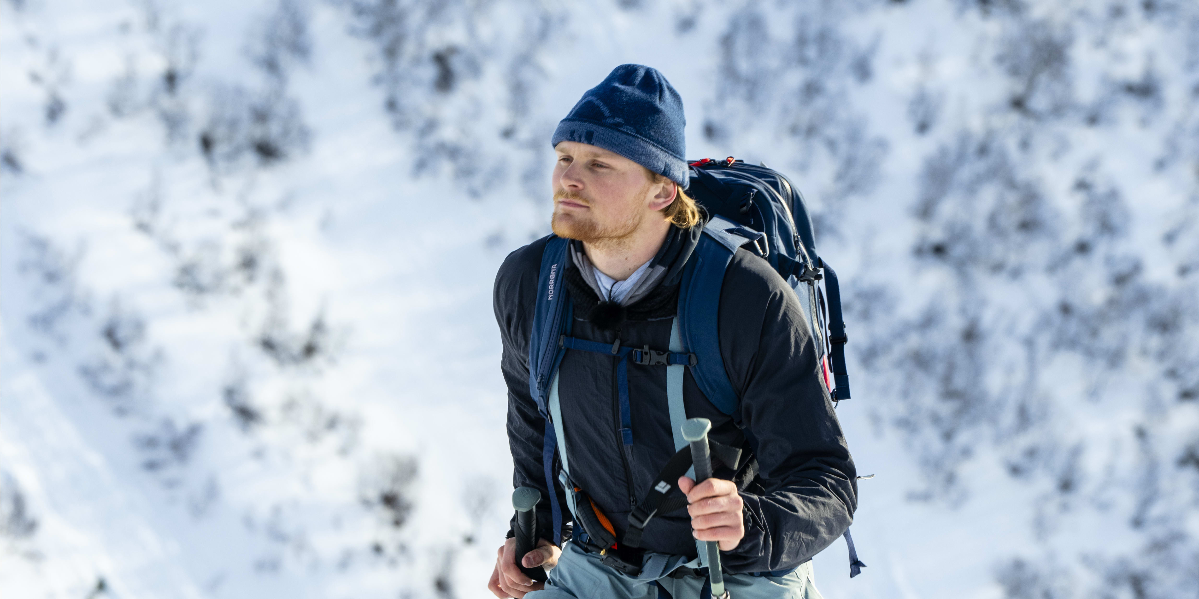 Man wearing Norrøna pants and a backpack, standing on skis in snowy mountains. 
