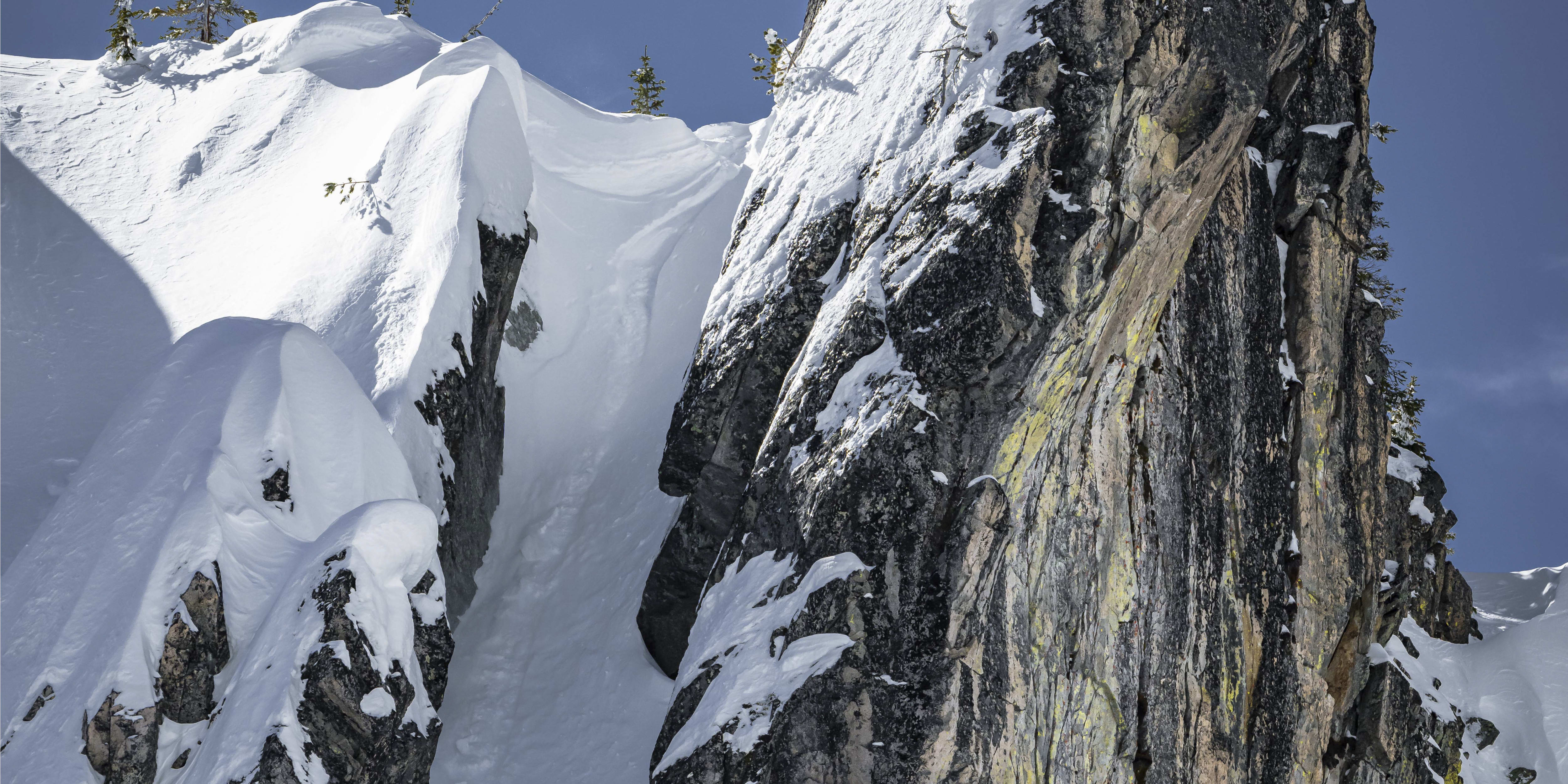 A person in a yellow jacket snowboards down a steep slope between two large, snow-covered rocks. The sky is blue.