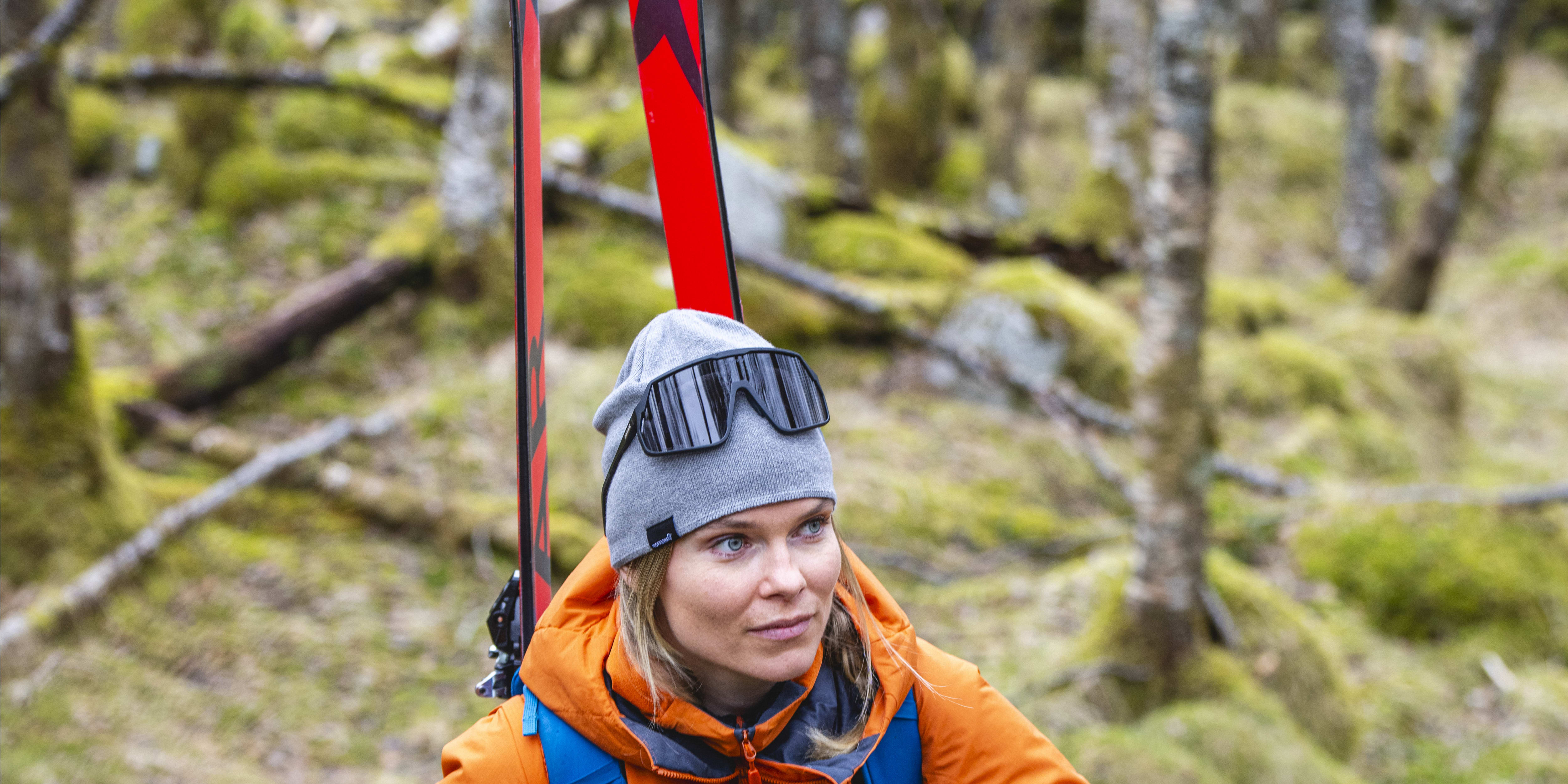 A woman hikes uphill through a wooded area carrying skis and poles. She wears a grey Norrøna beanie, goggles, and an orange Norrøna jacket with a blue backpack.