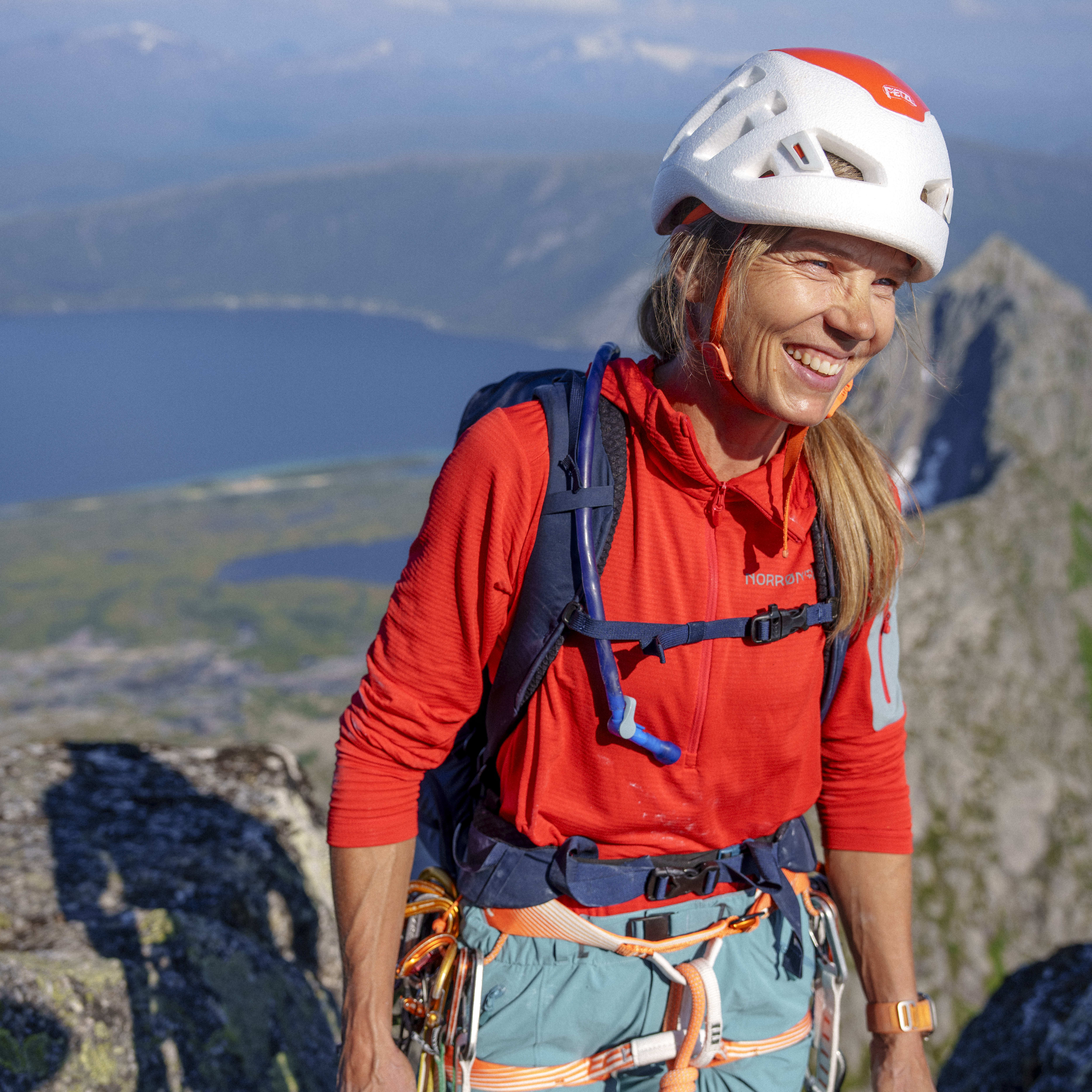 A climber smiles on a mountaintop, wearing a helmet, red Norrøna hoodie, blue pants, and climbing gear. Distant mountains and a lake are visible.