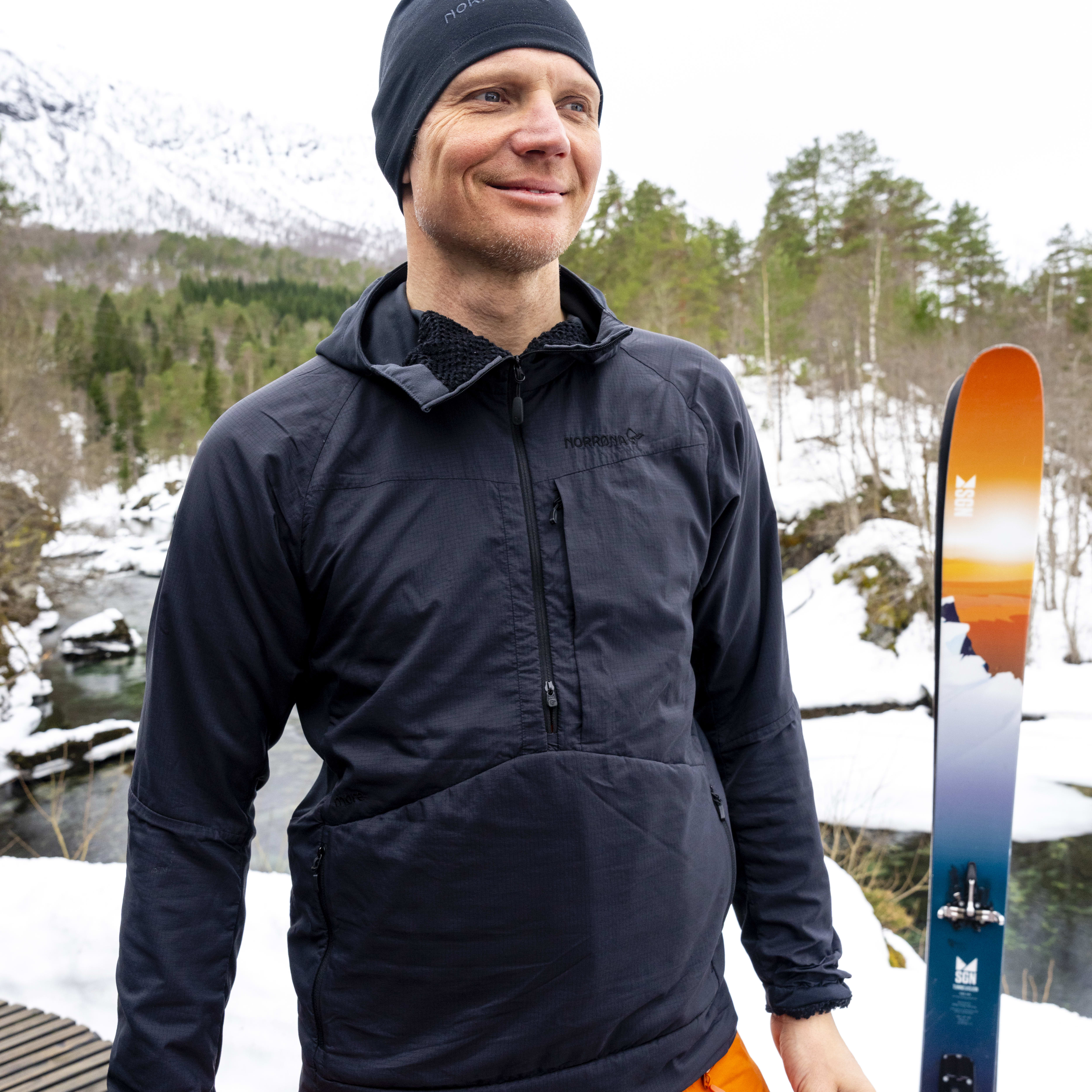 A man wearing a Norrøna jacket and orange pants stands on a wooden platform with a pair of skis in the snow.  
