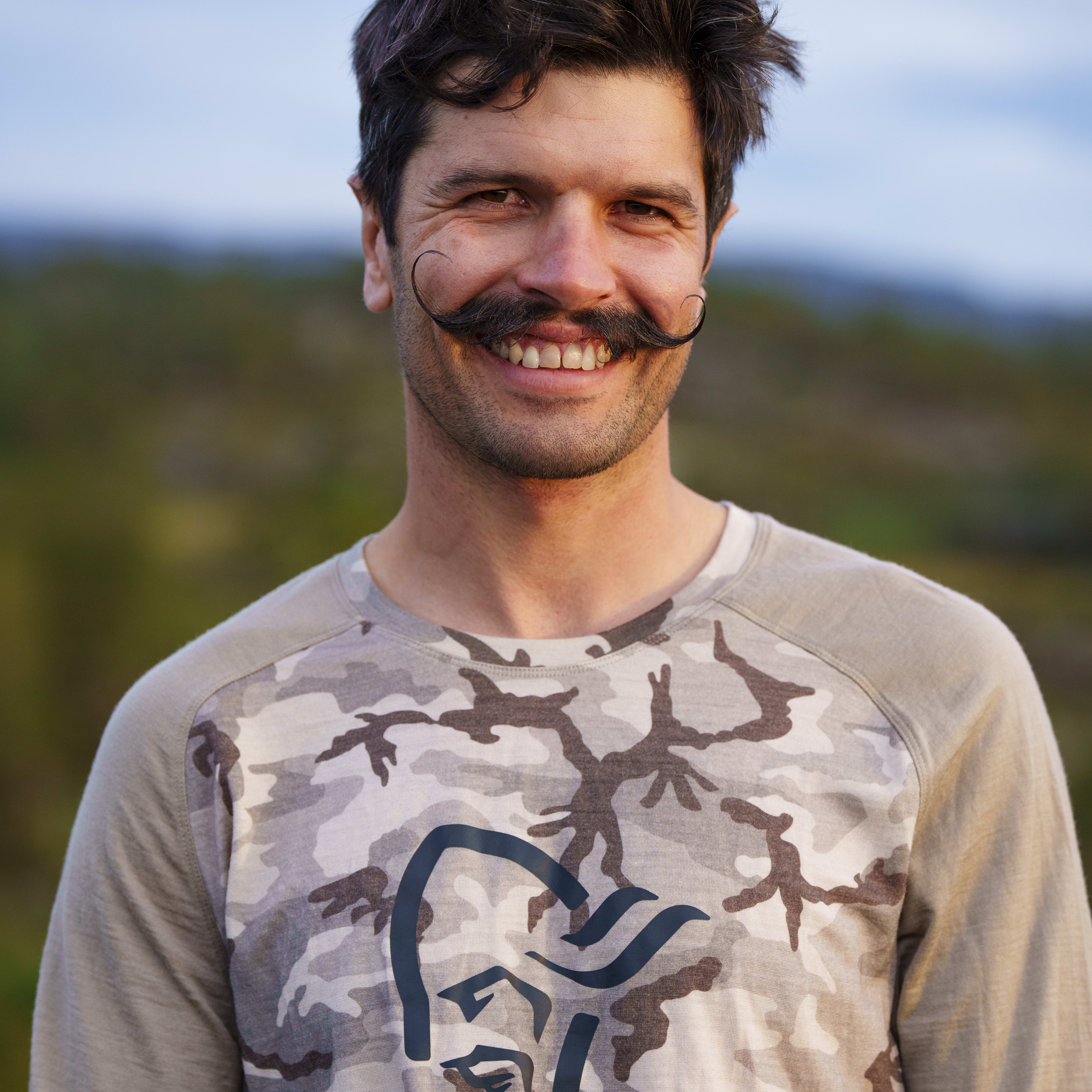 A man with a mustache is smiling. He is wearing a Norrøna camo shirt. 
