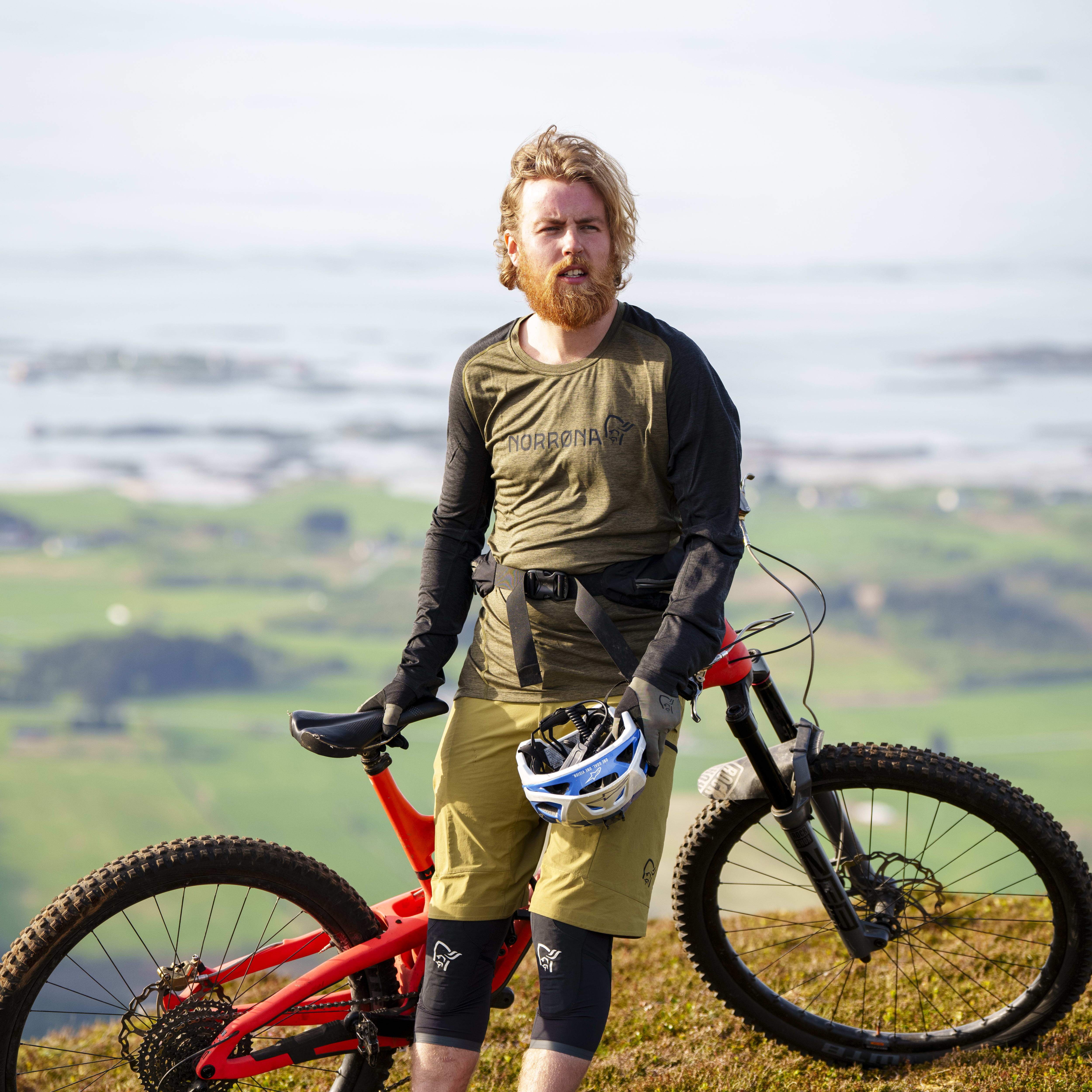 A man with a red beard stands next to his mountain bike on a grassy hillside. He is wearing a green long-sleeved shirt with black sleeves, tan shorts, knee pads, and black shoes. He is holding a white helmet in his left hand. The background is blurred and shows a body of water and more hills.
