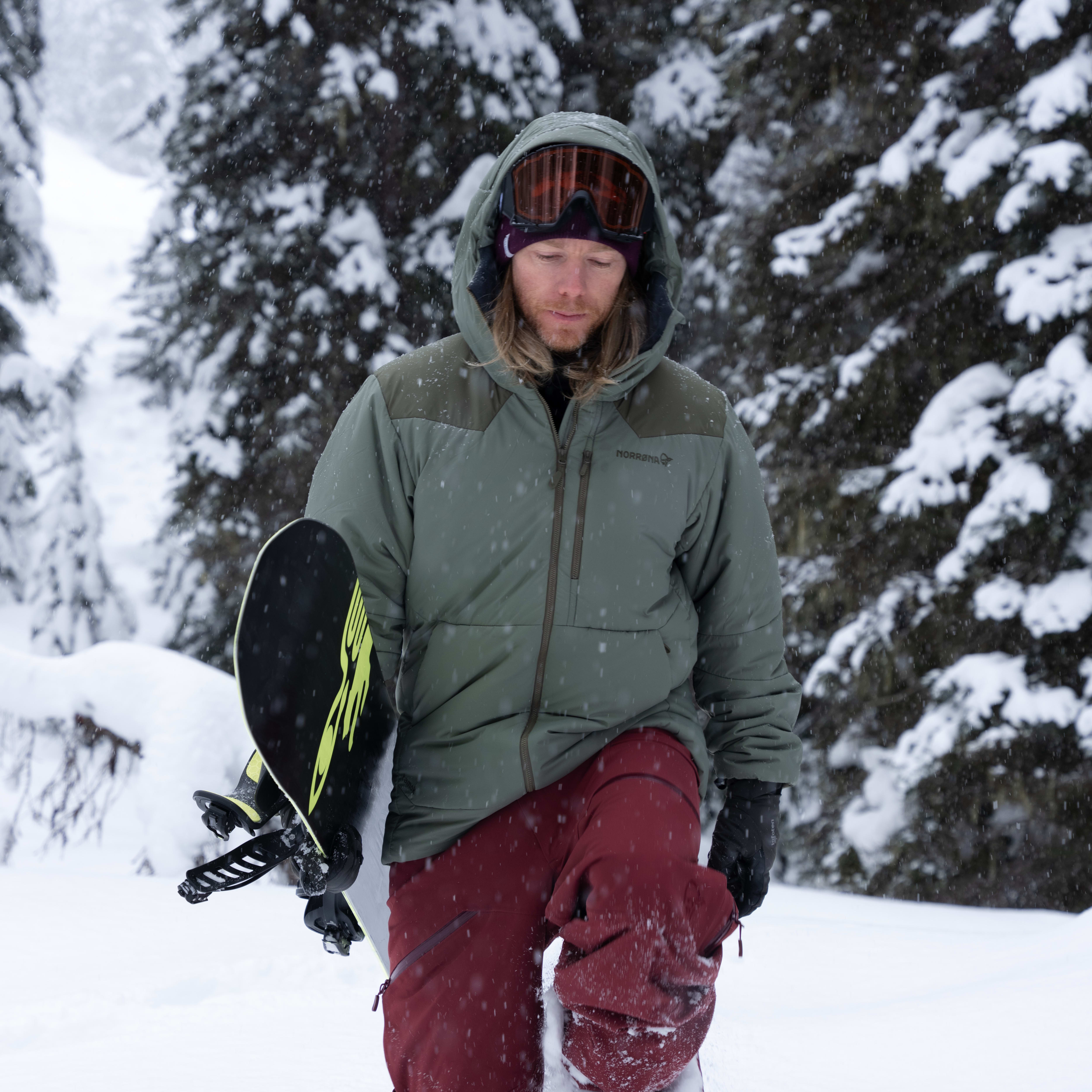 A man wearing a Norrøna jacket and pants, goggles, and carrying a snowboard walks in the snow. Snowy trees in background.