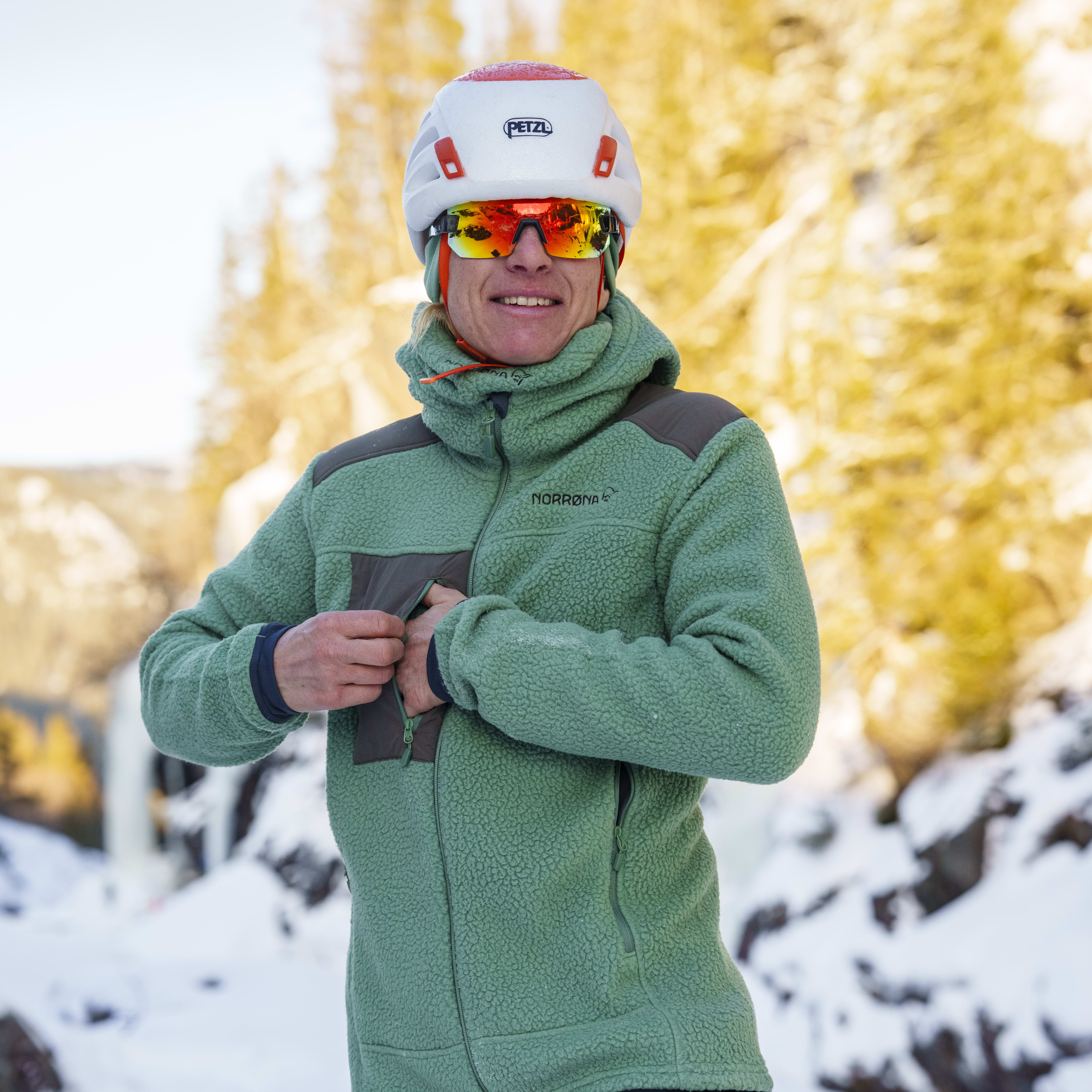 A man wearing a green Norrøna fleece jacket and black Norrøna pants, a white helmet with red accents and goggles, standing in snow.