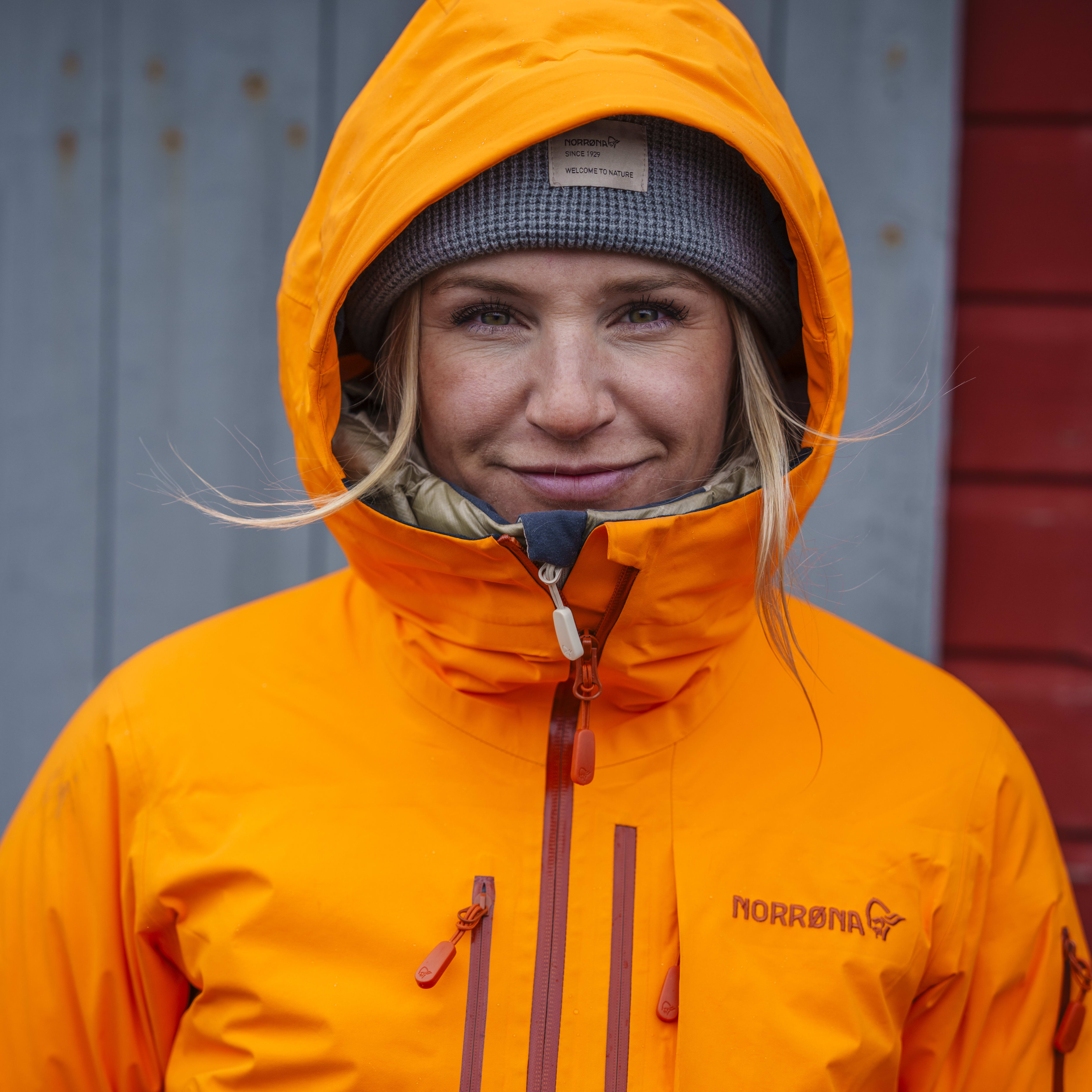 A woman wearing an orange hooded jacket, a grey knitted beanie, and looking directly at the camera. The text on the jacket reads "Norrøna" and "Welcome to Nature" on the beanie.