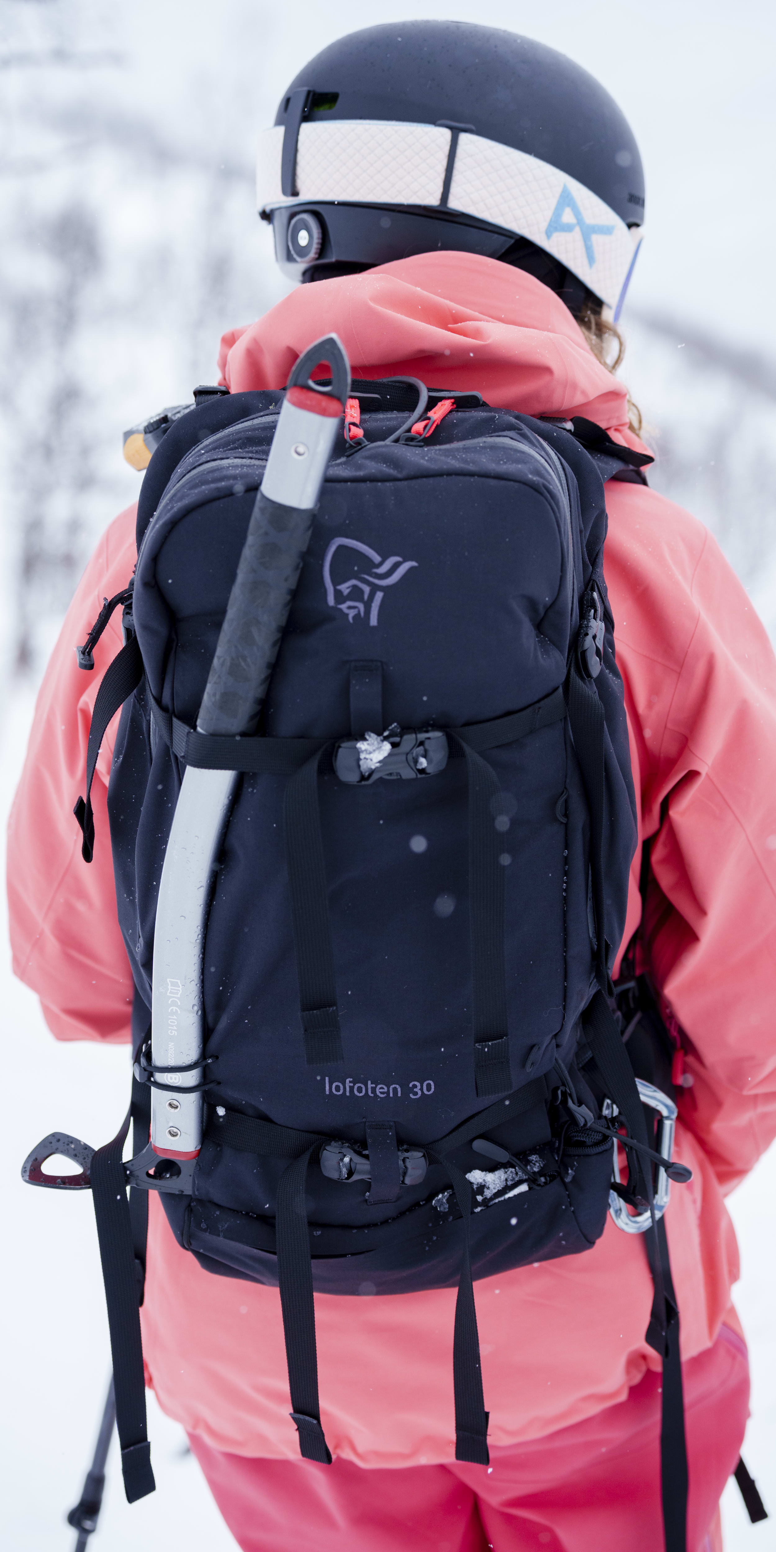 Back view of a skier in the snow wearing a helmet, pink Norrøna jacket, and a black 'Lofoten 30' backpack with an ice ax attached.