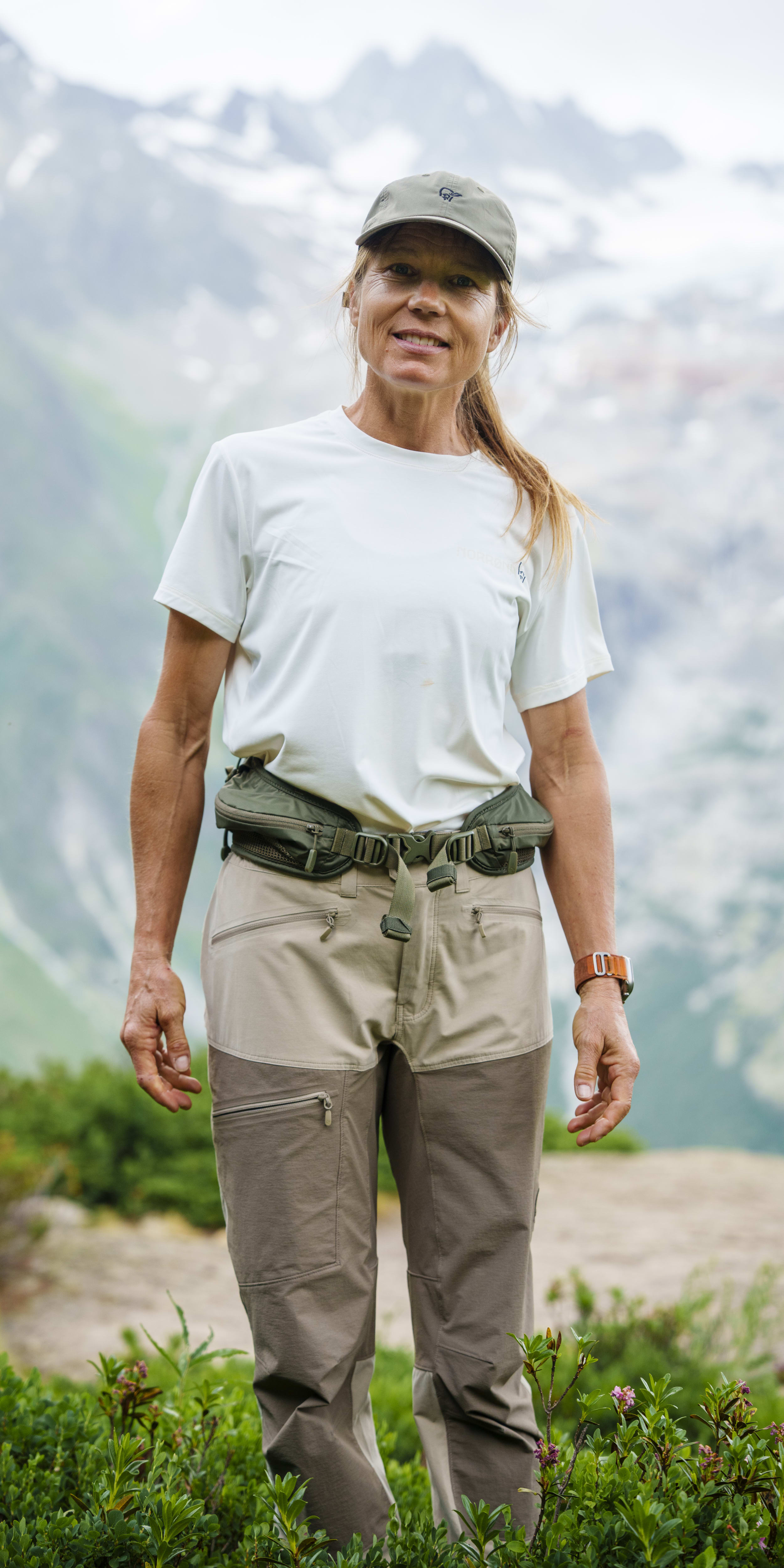 A smiling woman stands in front of a mountain range wearing a Norrøna cap and pants.