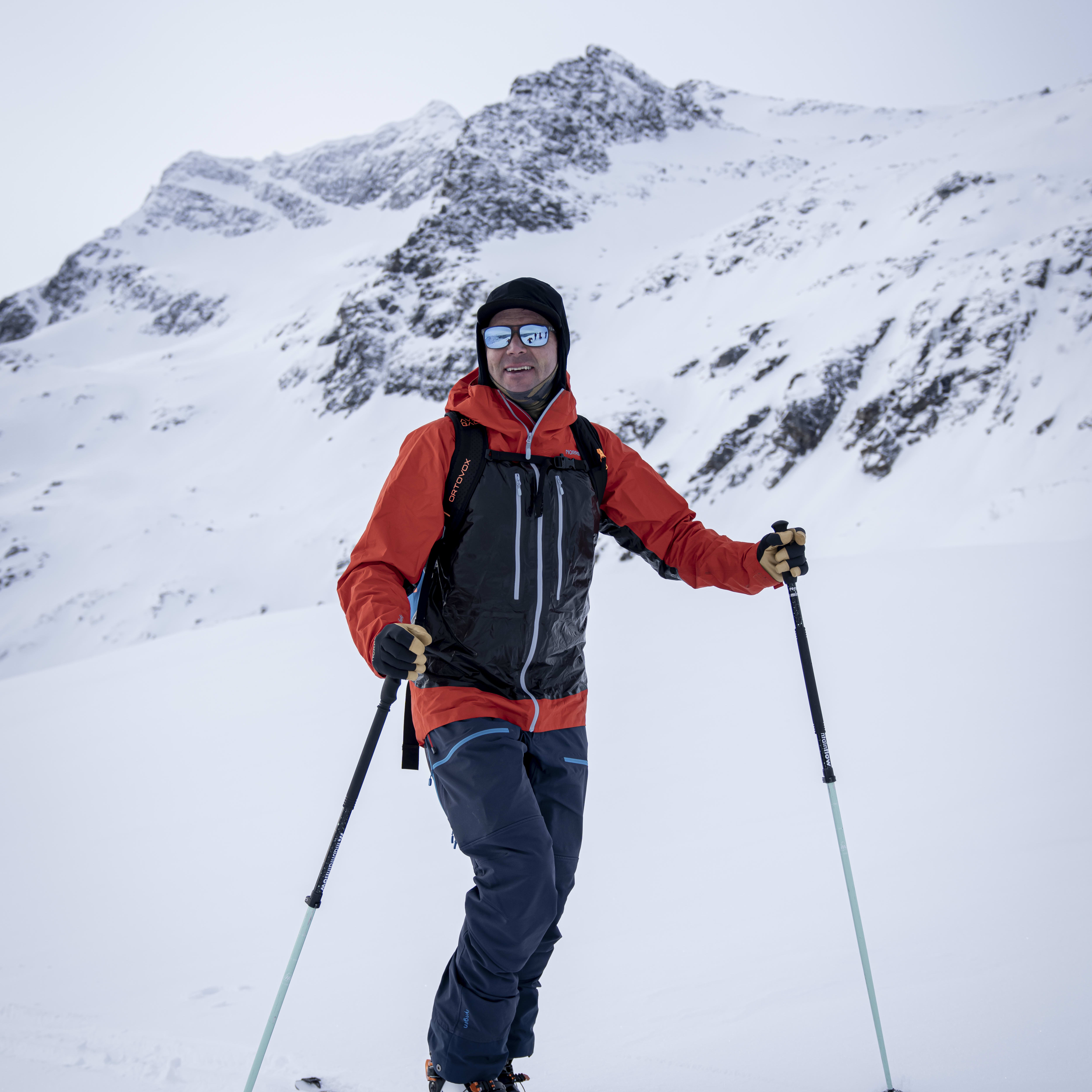 A skier in a Norrøna jacket and pants poses in snow with mountain backdrop. He is wearing skis and holding ski poles.