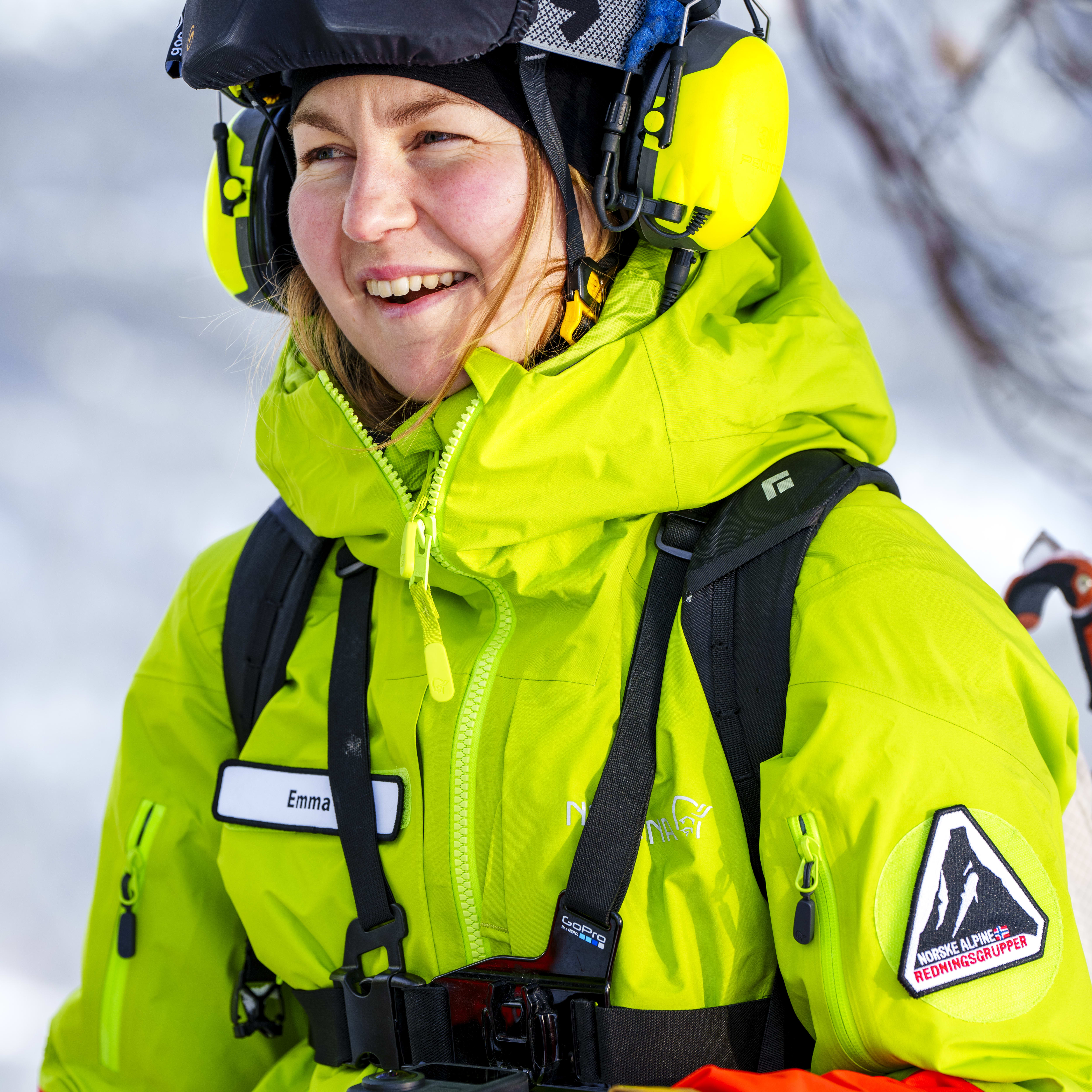 A woman wearing a yellow Norrøna jacket, a helmet, and ear protection, smiles at the camera. 
