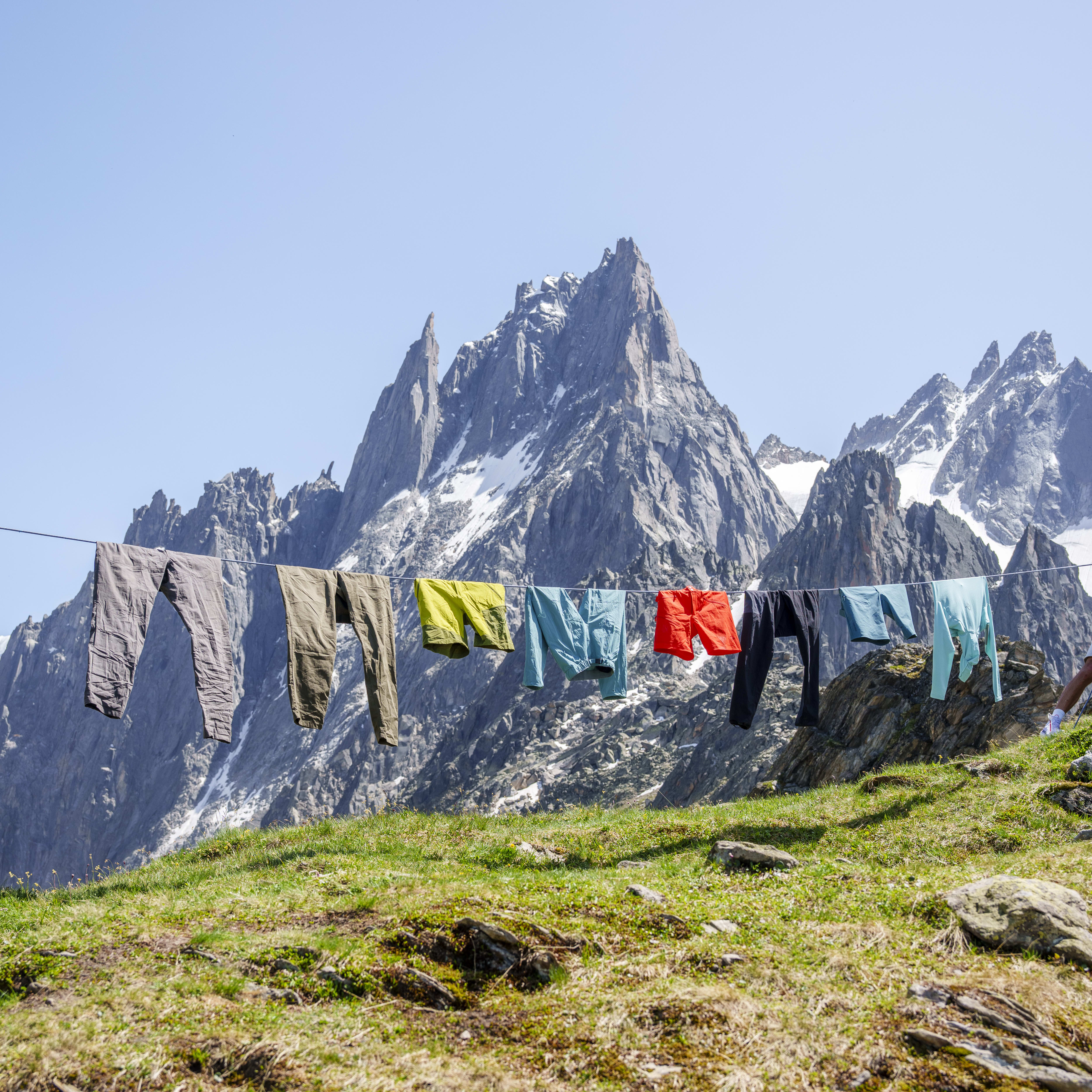 Norrøna hiking clothes dry on a line in an alpine meadow, with snow-capped peaks in the background under a clear blue sky.