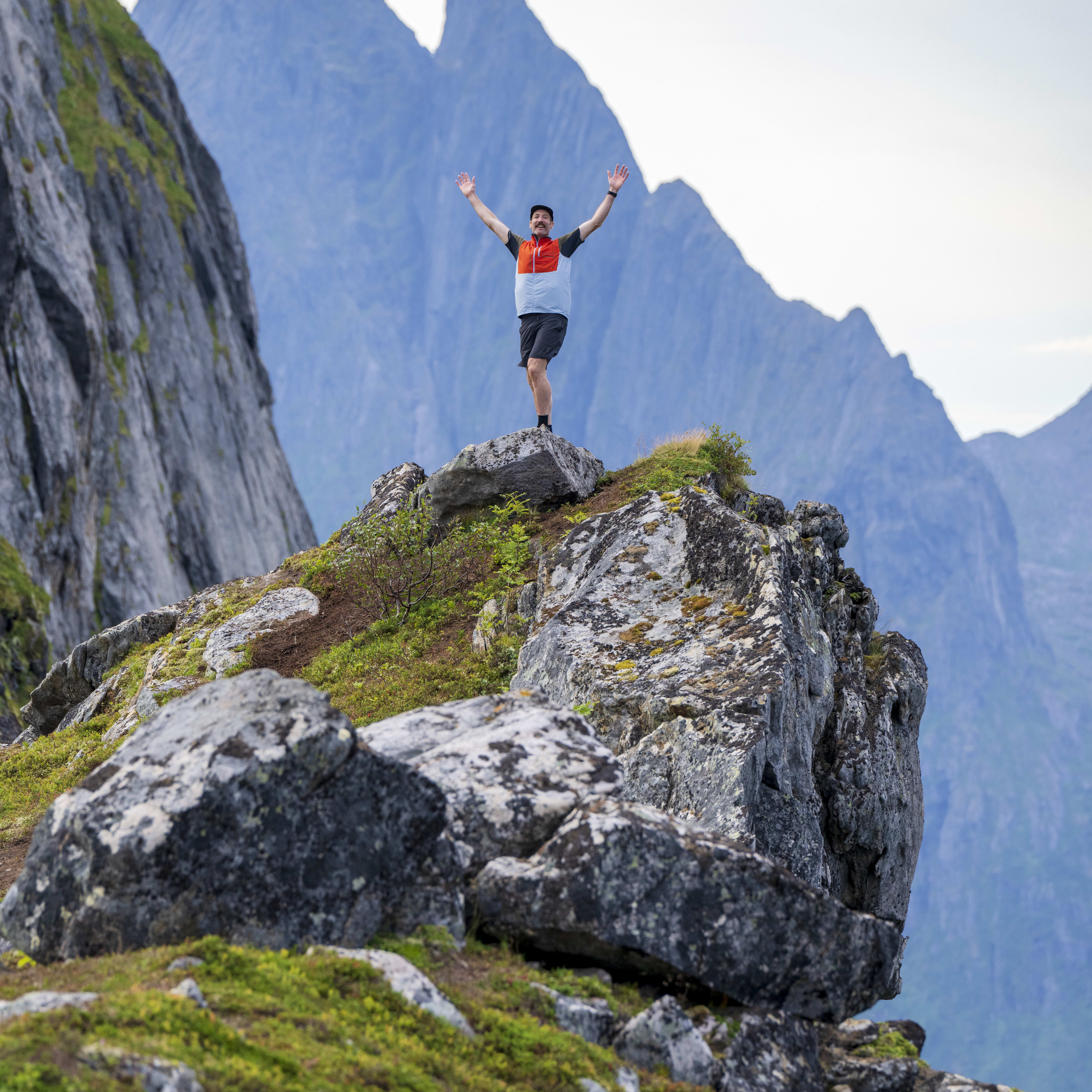 A man wearing a Norrøna vest stands on a rocky mountaintop with his arms raised.  
