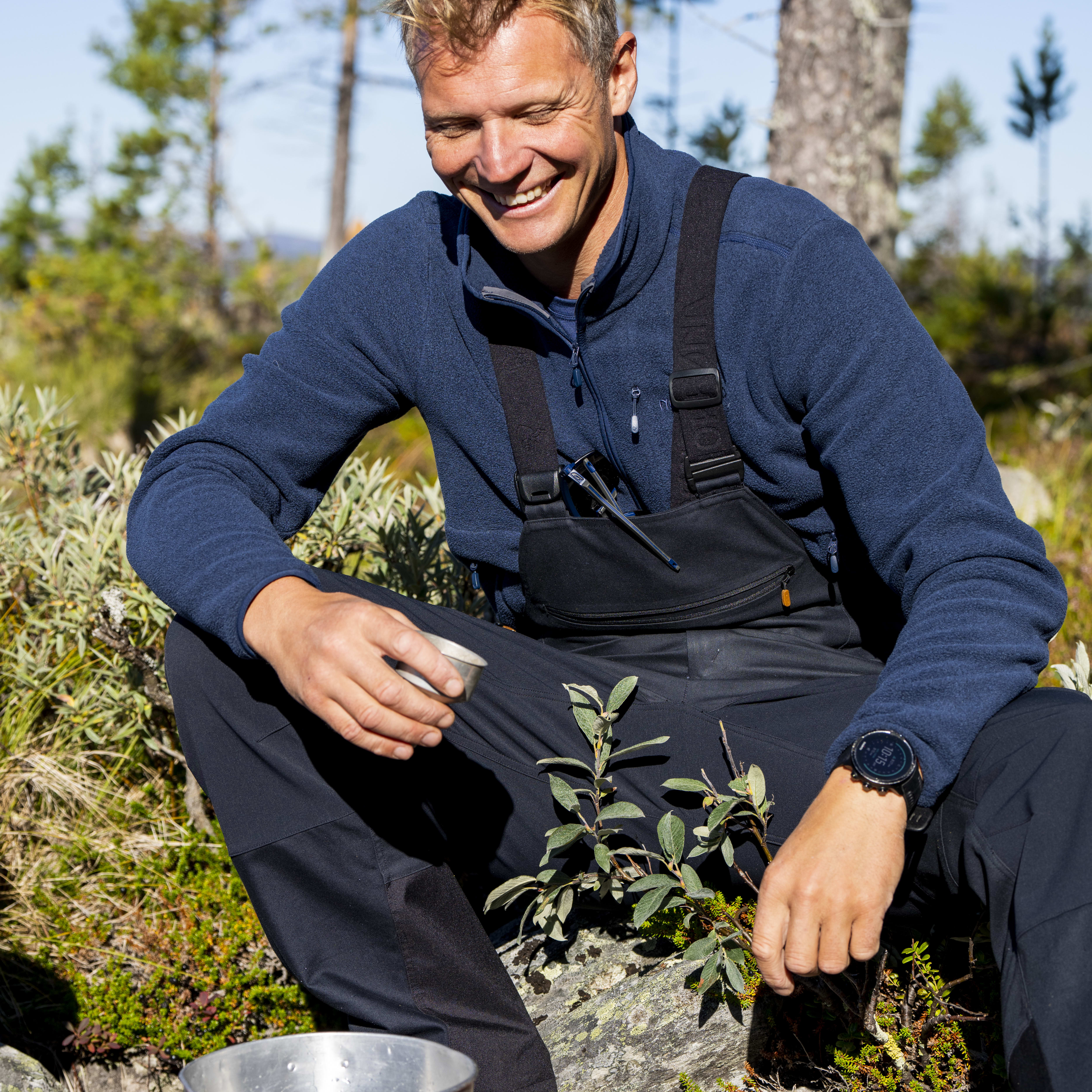 A man wearing Norrøna clothing sits on a rock, smiling while holding a cup, with a camping stove and pot in front of him.